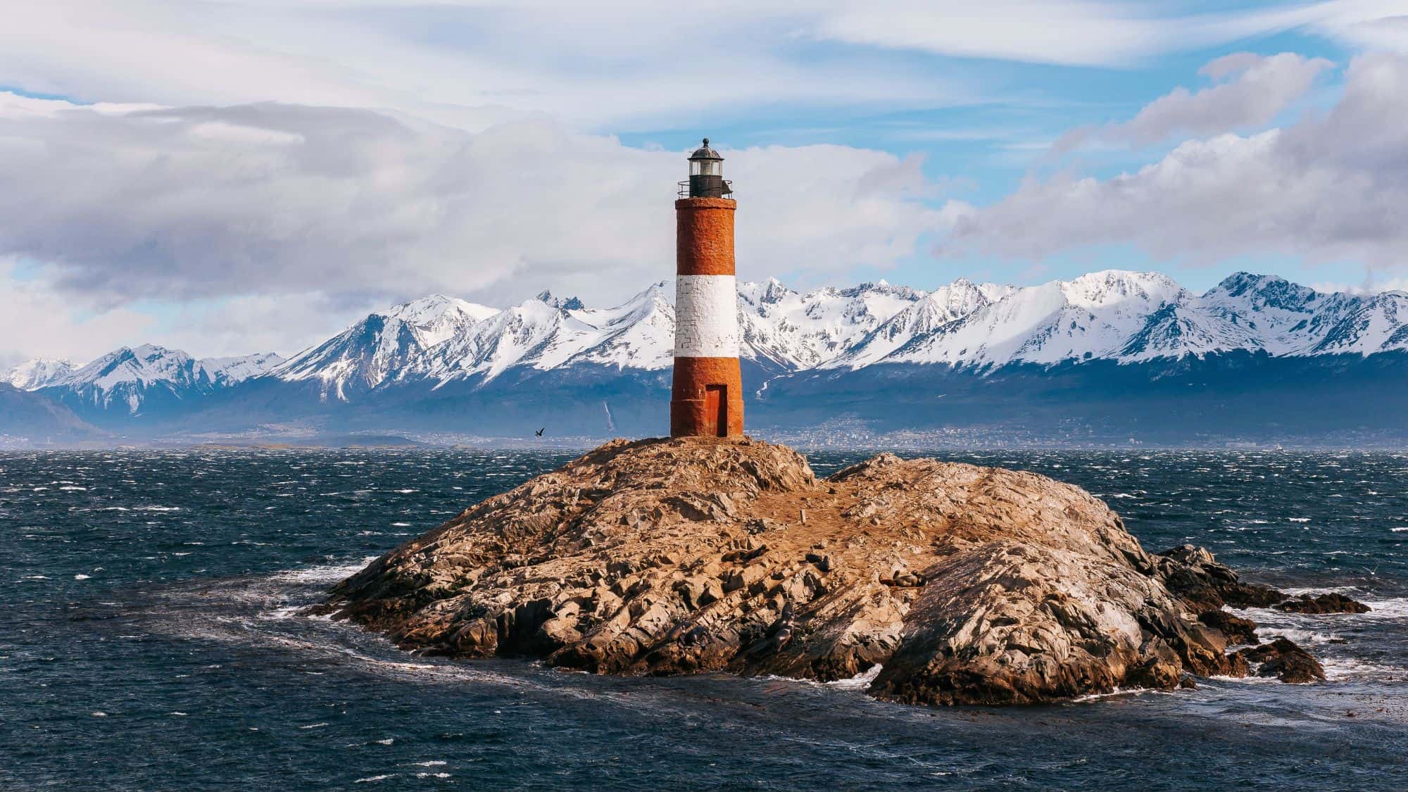 A red-and-white striped lighthouse stands on a rocky islet with snow-capped mountains in the distance.