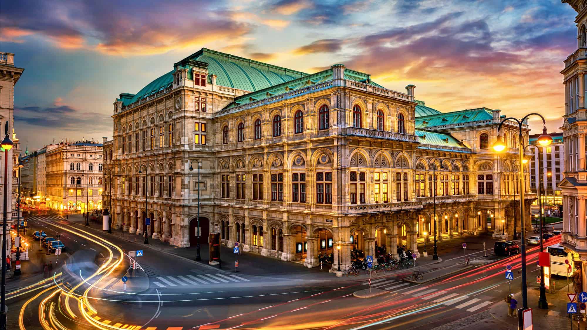 The illuminated Vienna State Opera glows at dusk as light trails from passing cars curve through the lively city streets.