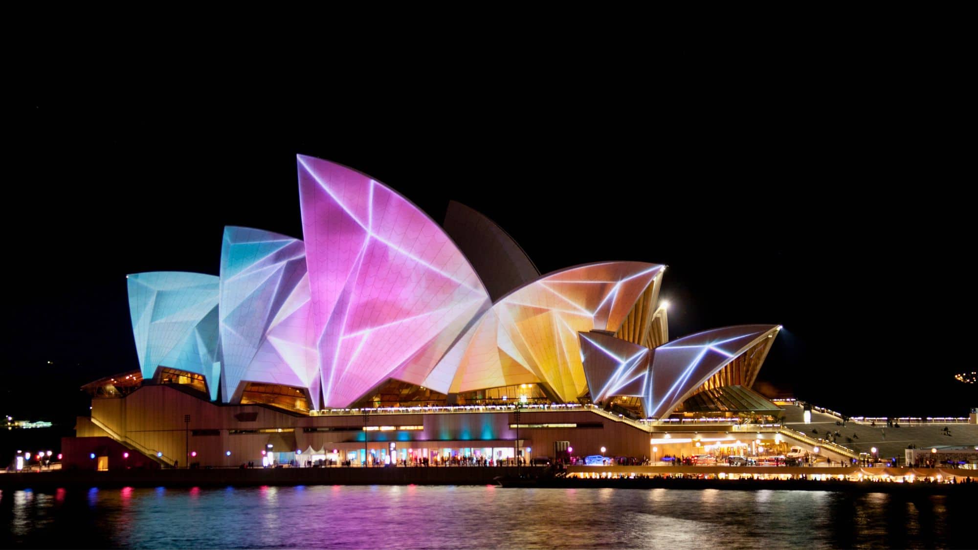 The Sydney Opera House sails are illuminated with pink, blue, and gold light projections during a nighttime festival.