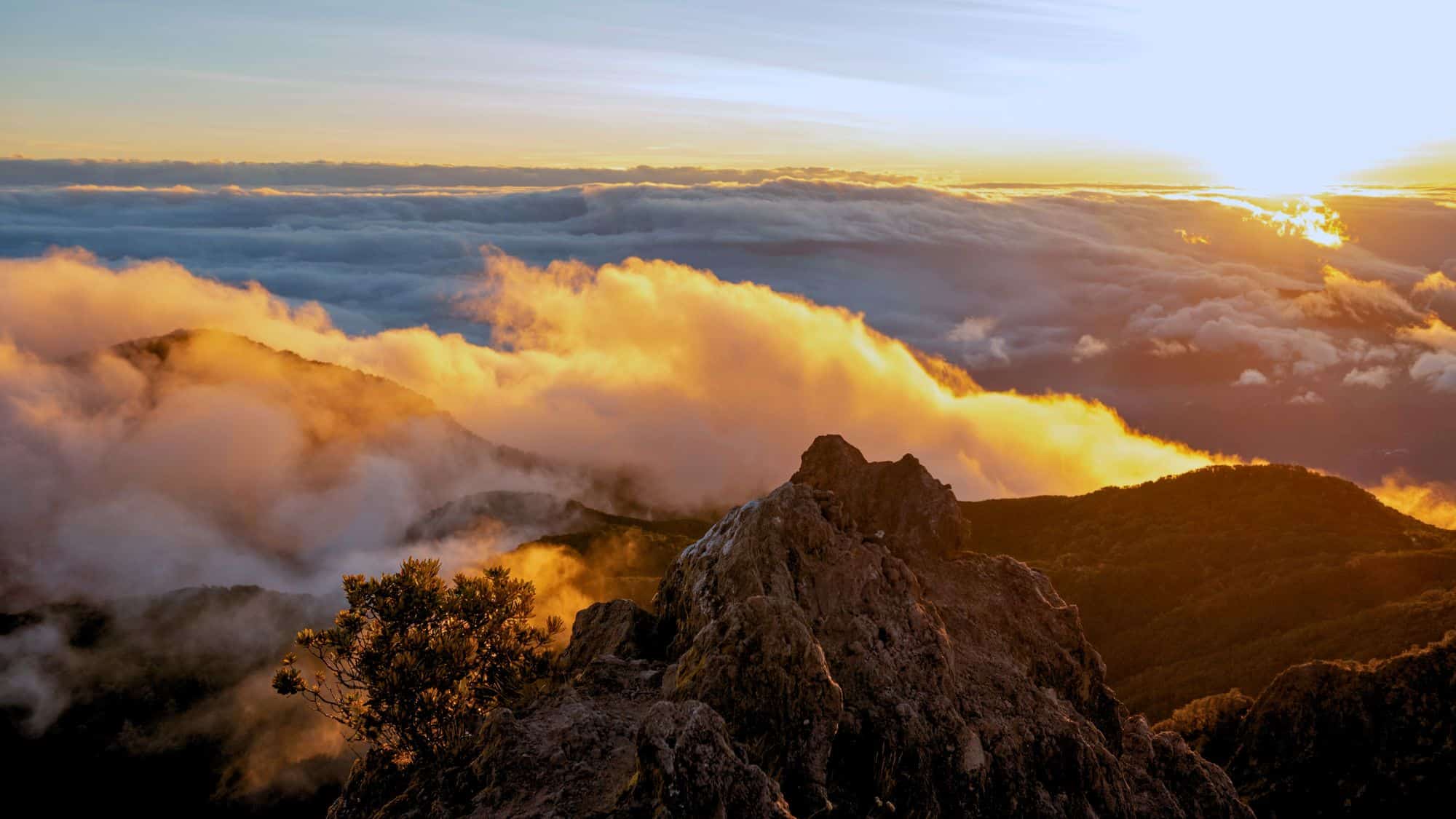 Fiery orange clouds roll over the lush mountain peaks of Volcán Barú at sunrise, with rocky outcrops and a glowing horizon above the mist.