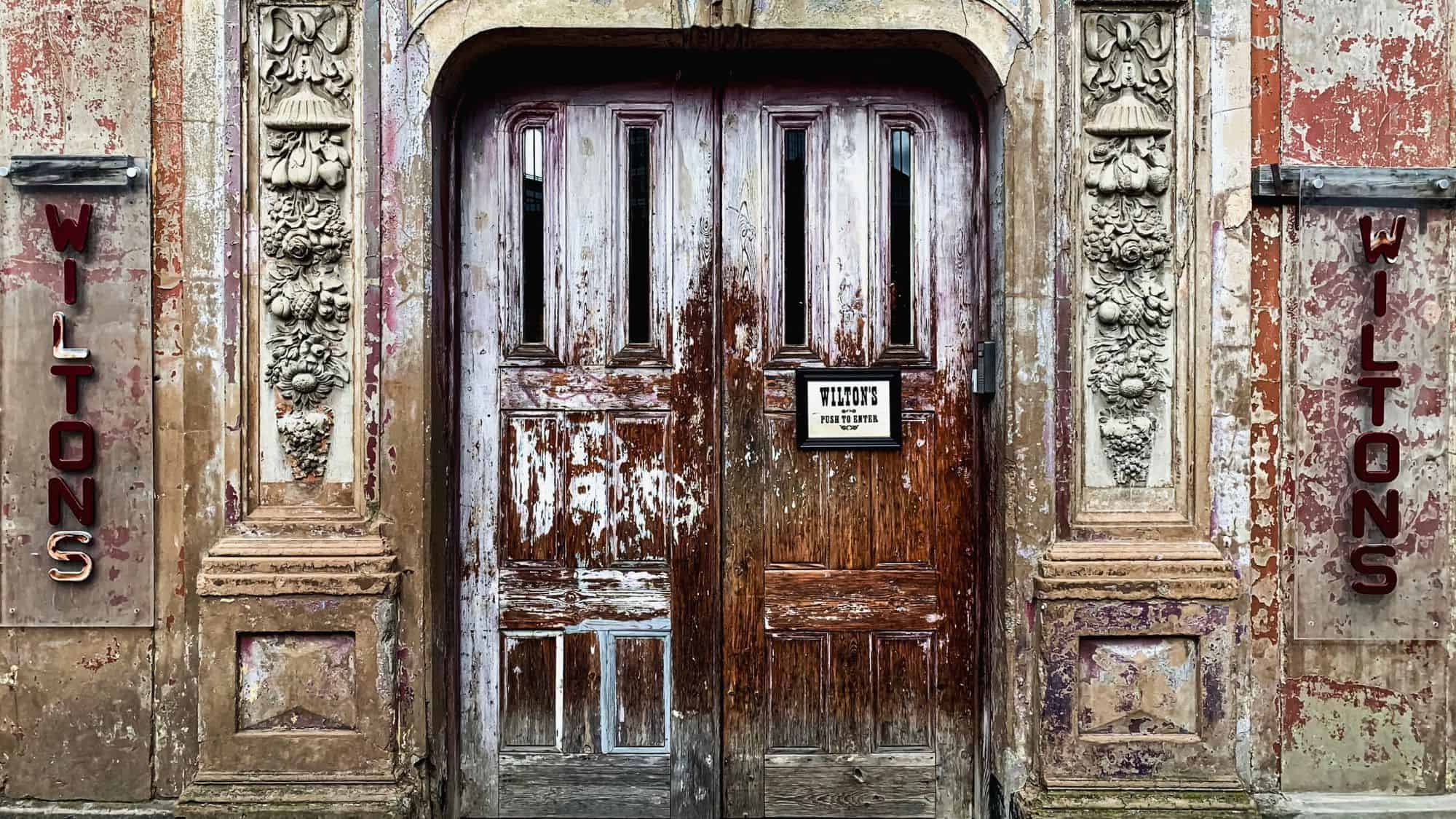 A weathered wooden door framed by ornate stone carvings marks the historic entrance to Wilton’s Music Hall.