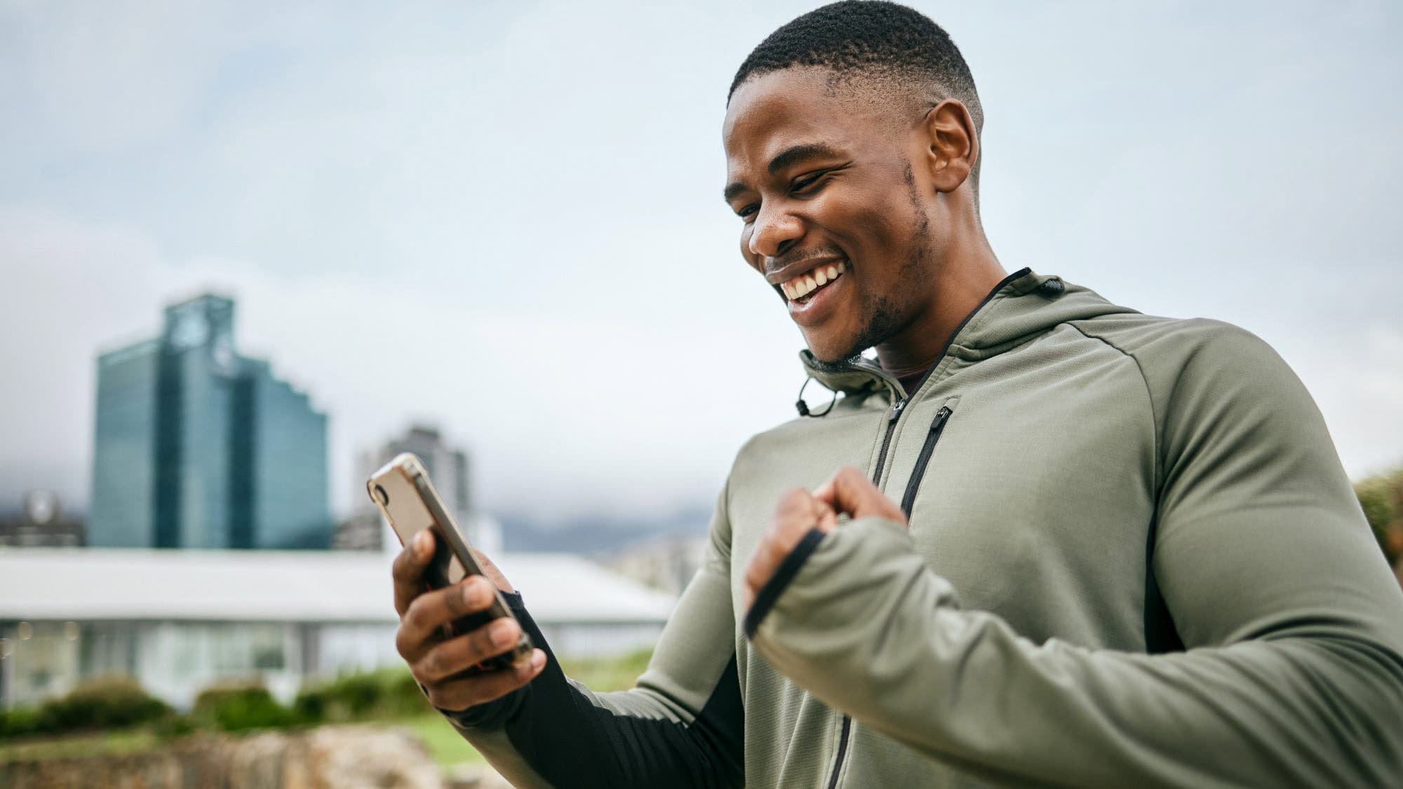 A man in athletic wear smiles and pumps his fist while looking at his phone, standing outdoors in front of a cityscape with tall buildings.