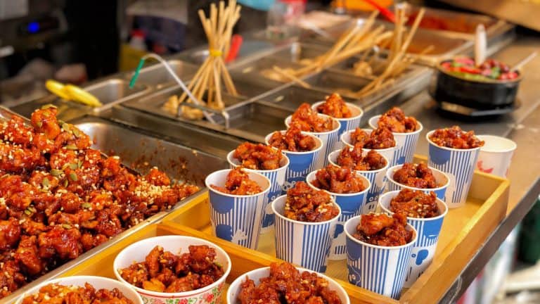 Crispy bite-sized fried chicken coated in a sticky, sweet-spicy glaze, topped with seeds, and served in striped paper cups at a street market.