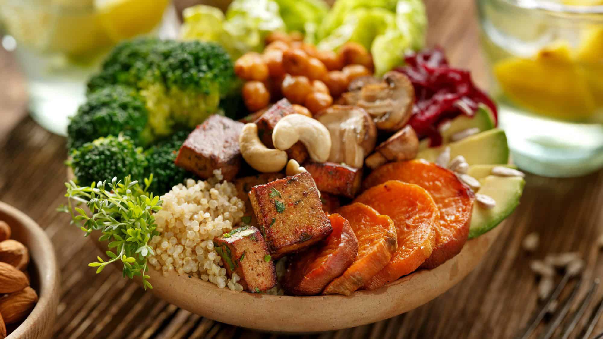 A wooden bowl is filled with a vibrant variety of healthy foods, including roasted sweet potatoes, tofu, quinoa, chickpeas, broccoli, avocado, and nuts.