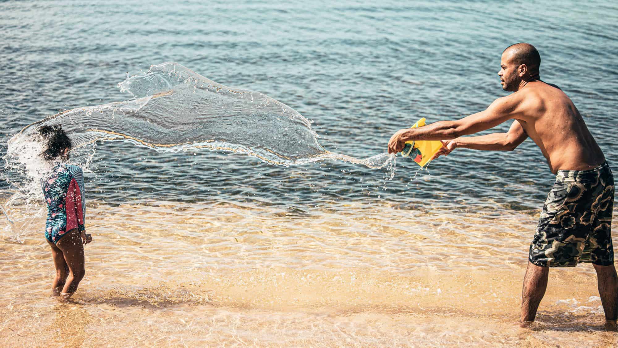 A man playfully tosses a bucket of water at a child standing at the shoreline, captured mid-splash.