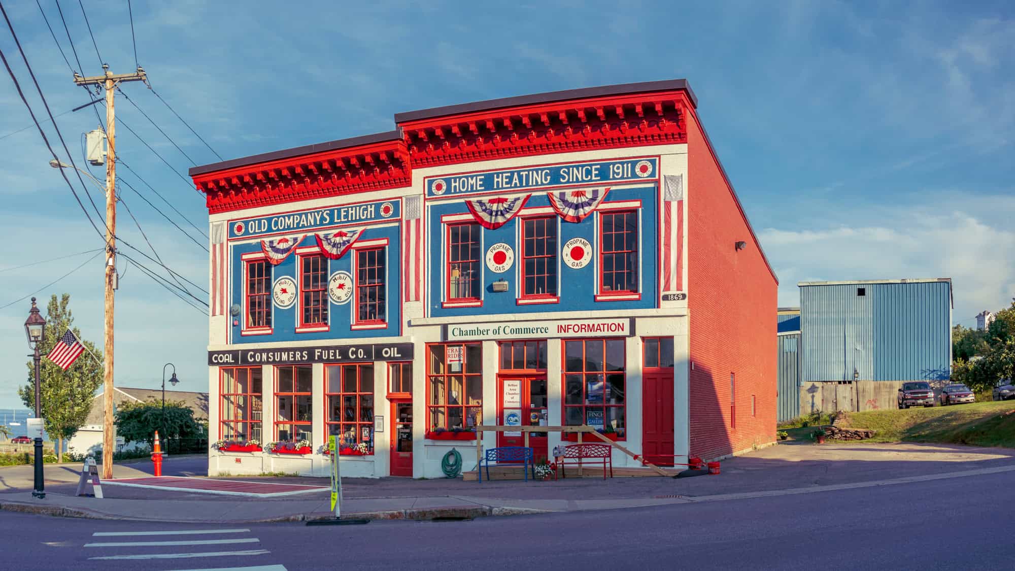 A bright red, white, and blue historic building displays signs for home heating and propane gas, decorated with bunting and American flags.