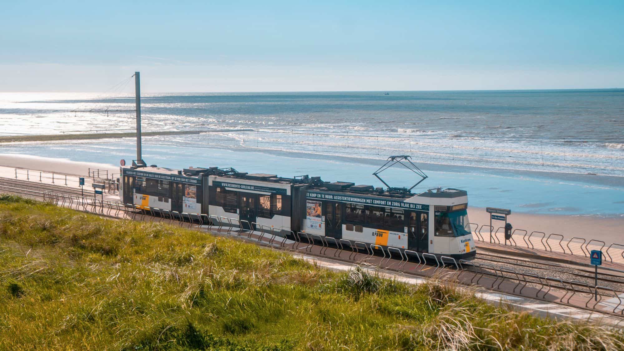 A seaside tram travels along tracks parallel to a sandy beach with waves rolling in under a bright blue sky.