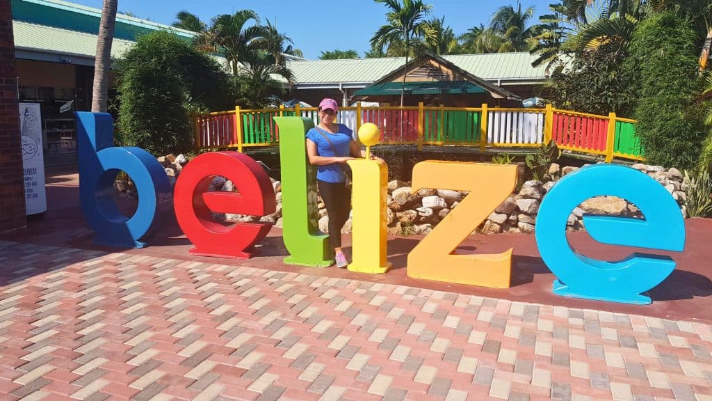 A person poses with a large, colorful “Belize” sign in a sunny plaza surrounded by palm trees and tropical plants.