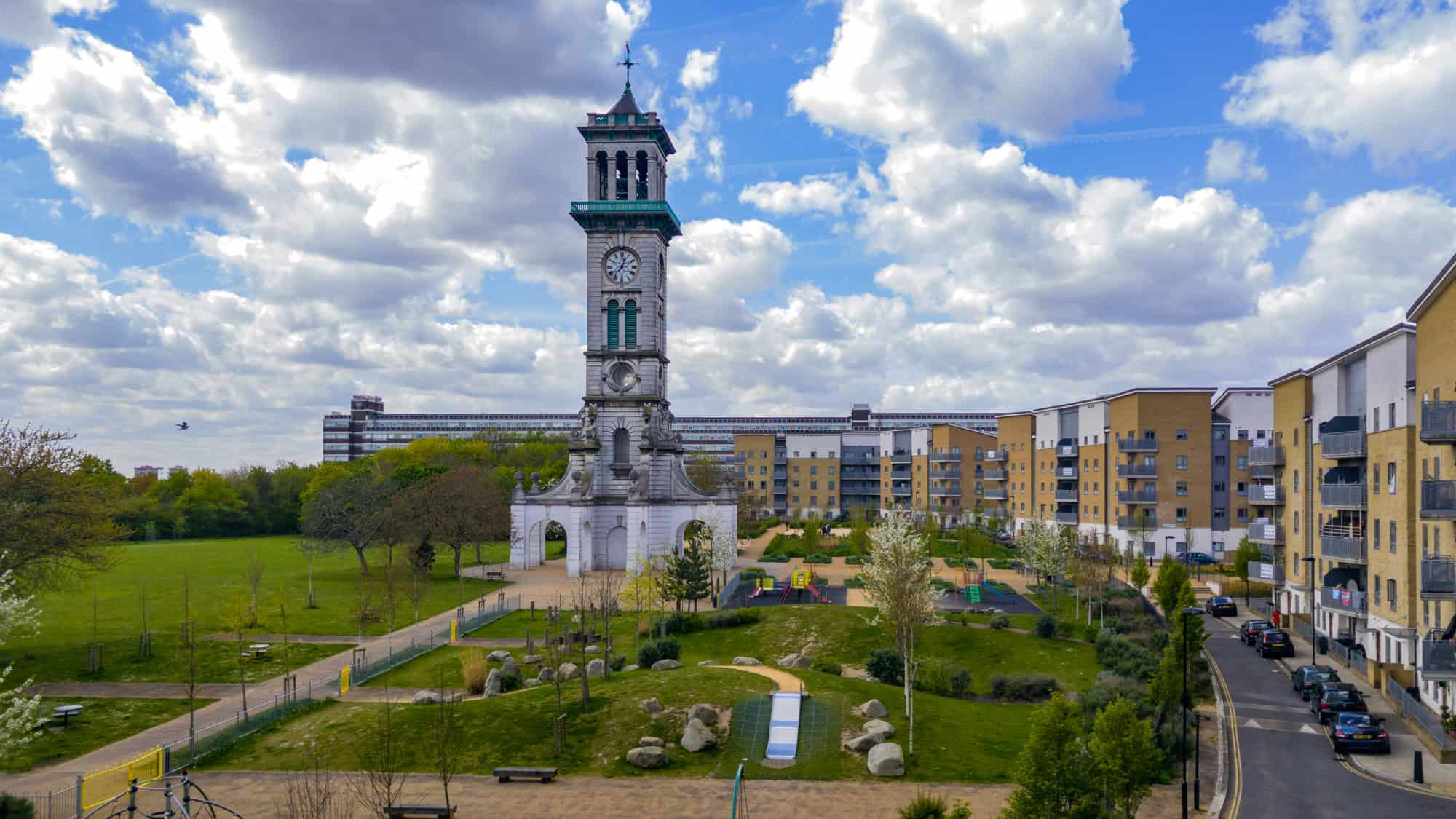 A tall stone clock tower rises in a grassy park surrounded by modern apartment buildings under a partly cloudy sky.