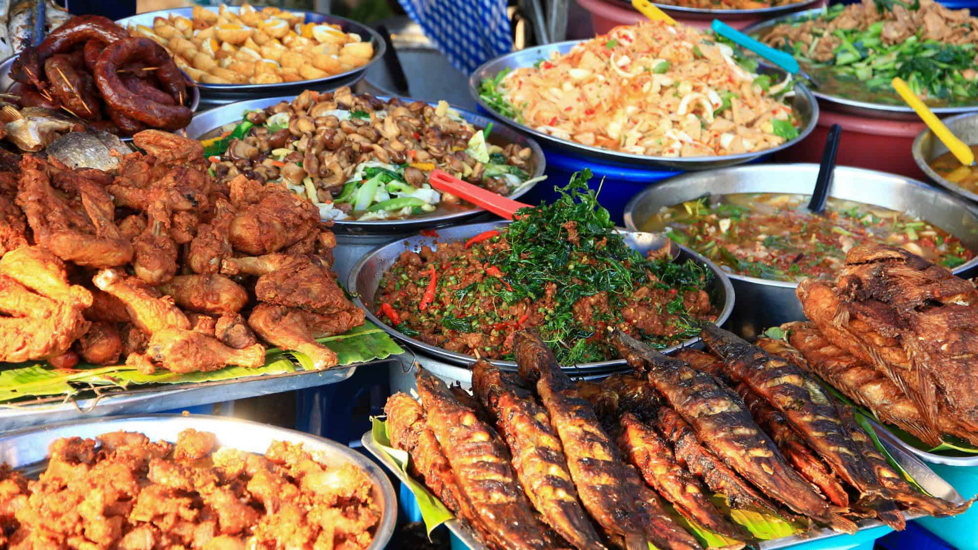 An abundant display of fried chicken, grilled fish, stir-fried vegetables, curries, and noodles in large metal trays at an outdoor market stall.