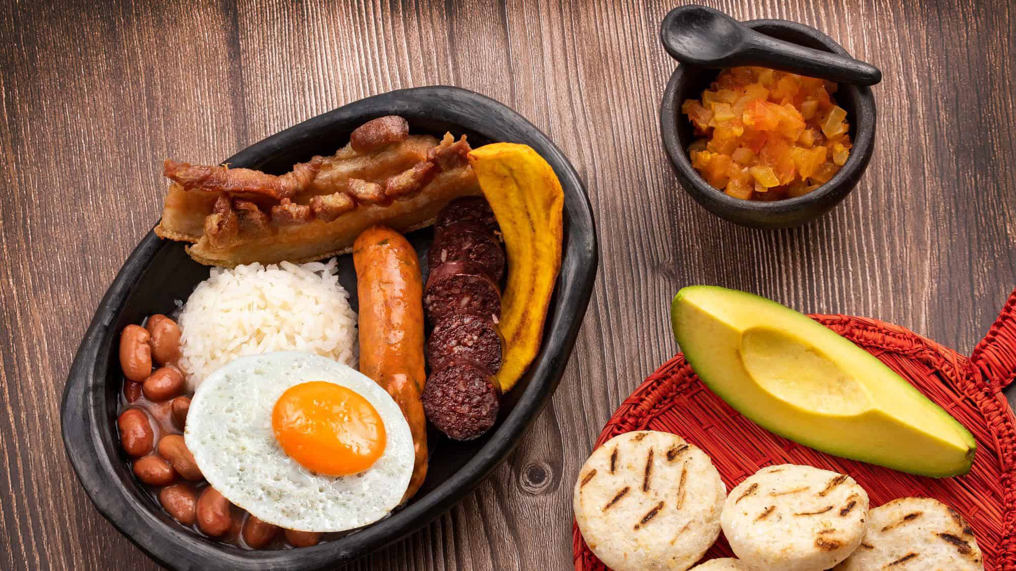 A traditional Colombian platter with rice, beans, fried egg, chorizo, morcilla, crispy pork belly, plantain, avocado, arepas, and a side of hogao sauce.