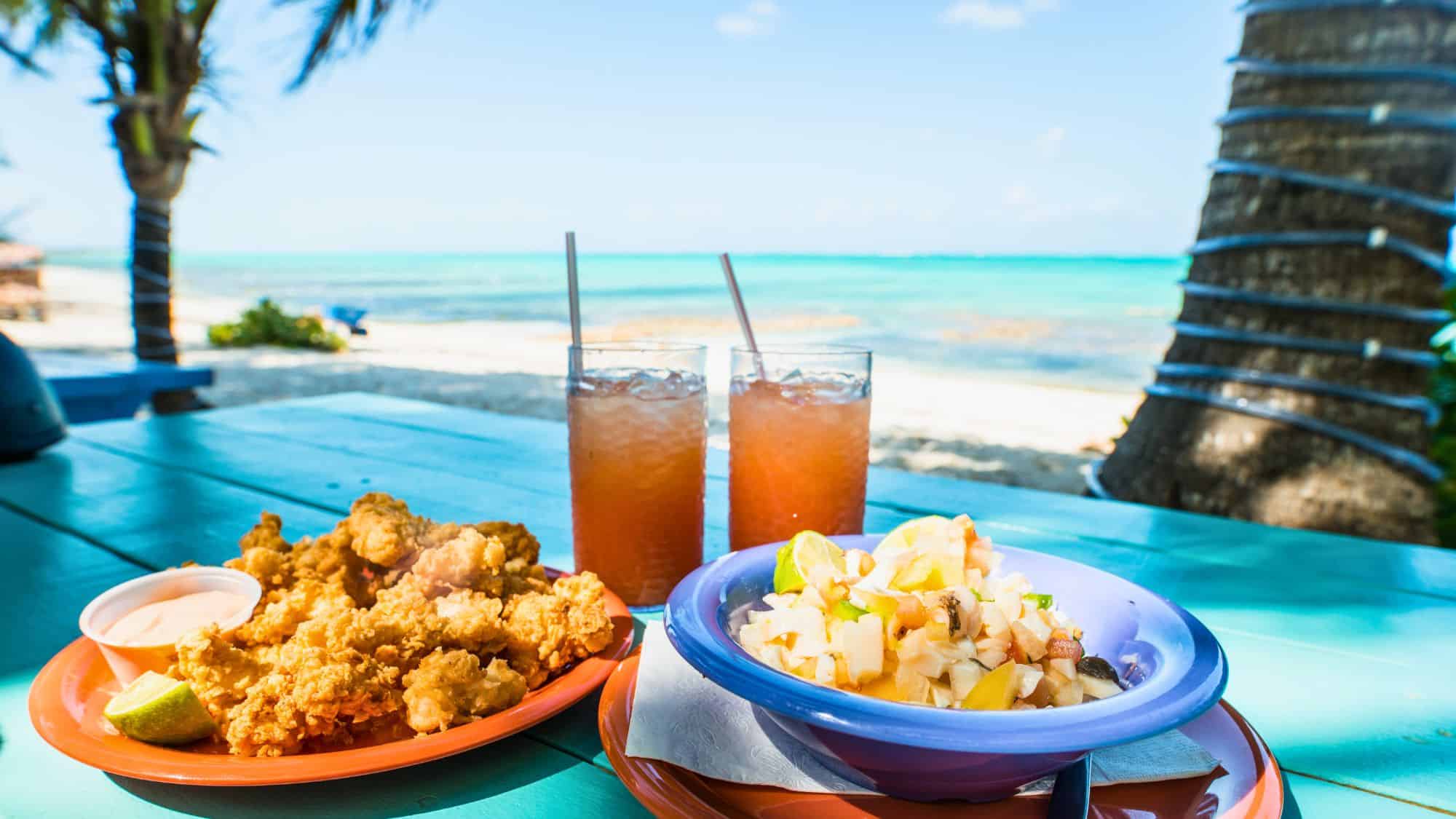 A colorful spread of fried seafood, ceviche, and two iced drinks sits on a turquoise picnic table with a view of the ocean and palm trees in the background.