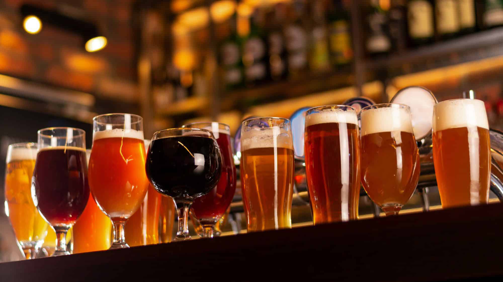 Glasses of beer in various shades from pale to dark are lined up on a bar counter in a pub.