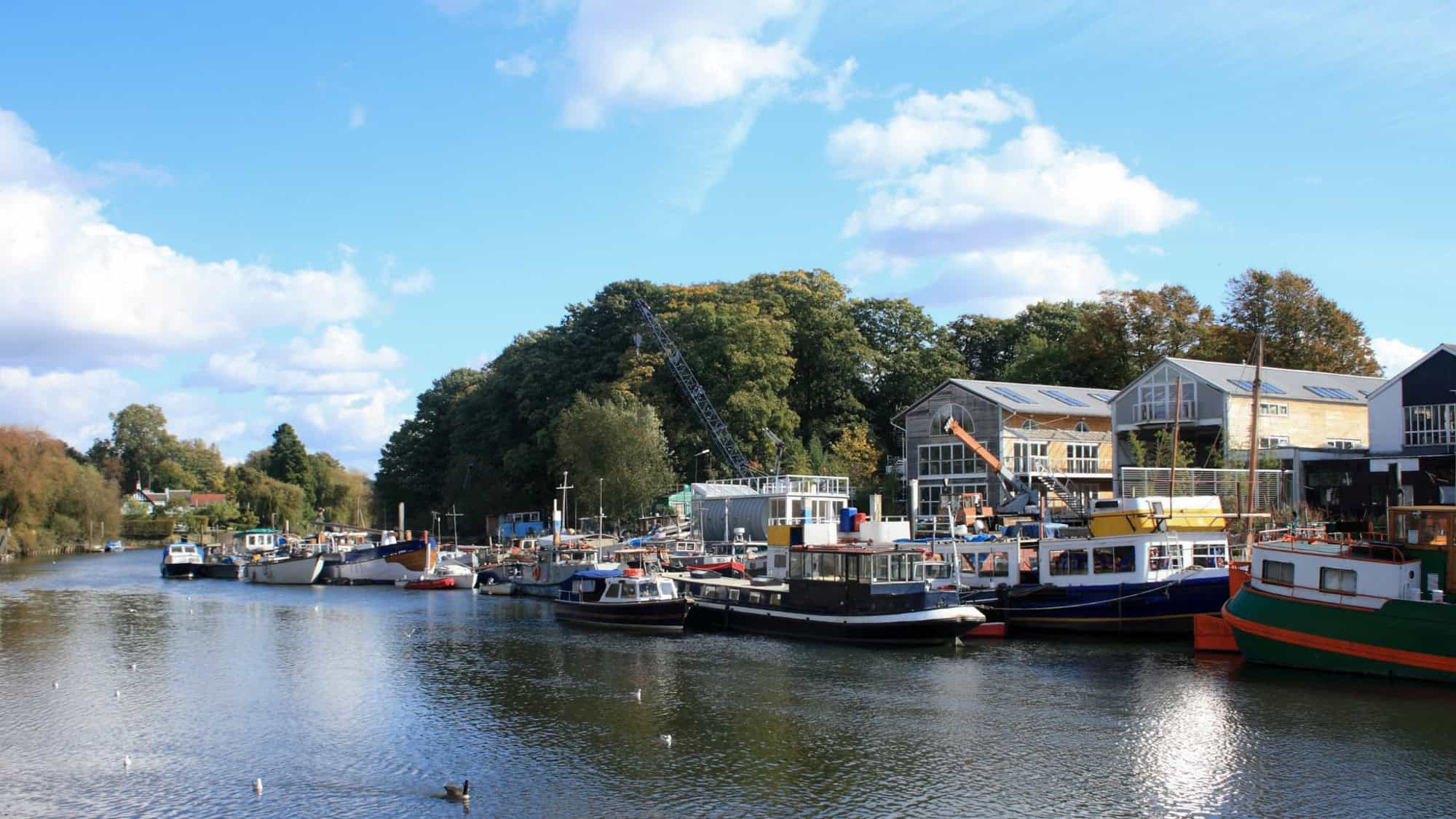 A cluster of boats and houseboats line a calm river beside warehouses and greenery under a blue sky.