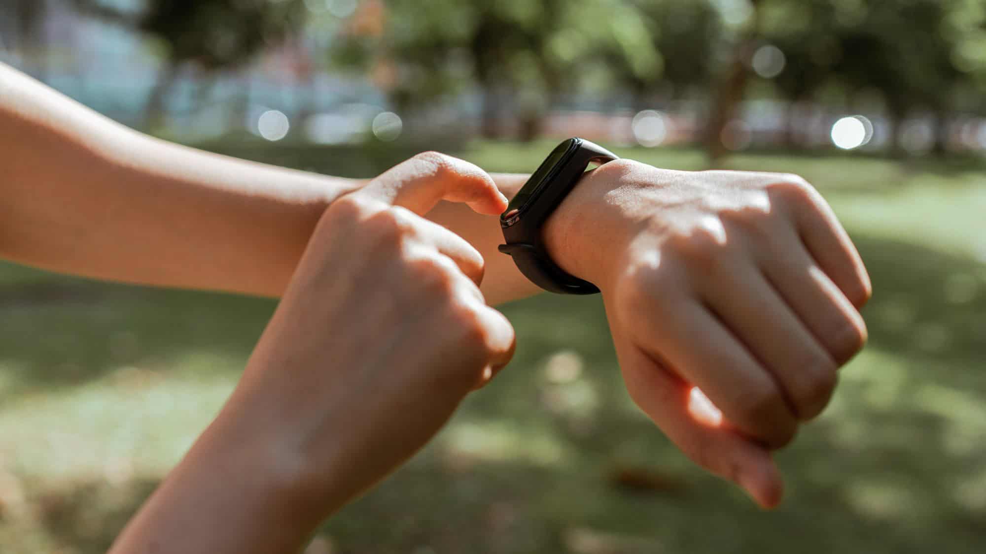 A close-up of hands shows someone interacting with a black fitness tracker or smartwatch in a sunny park with blurred greenery in the background.