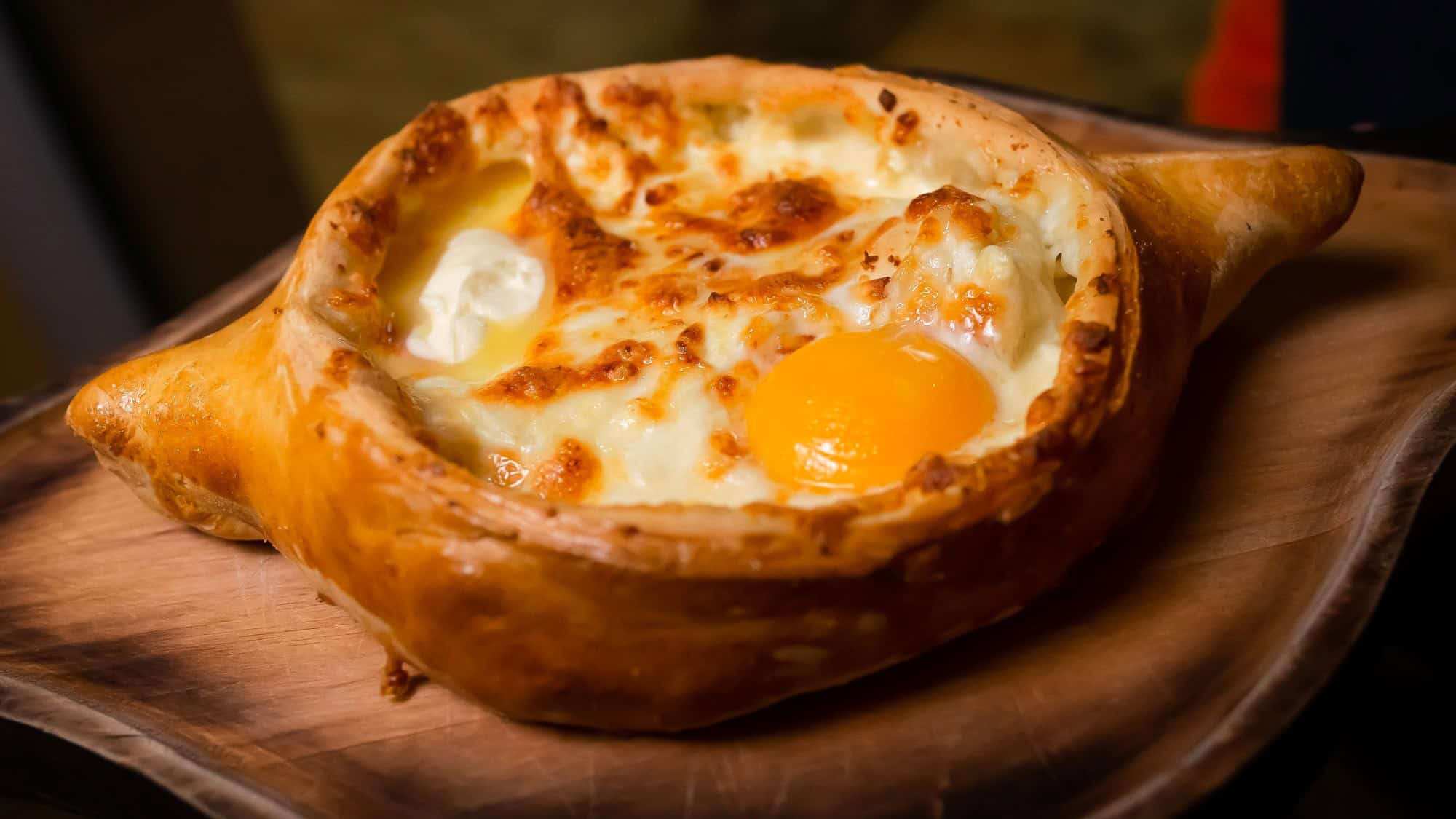 A golden boat-shaped bread filled with melted cheese, a runny egg yolk, and butter, served on a wooden board.