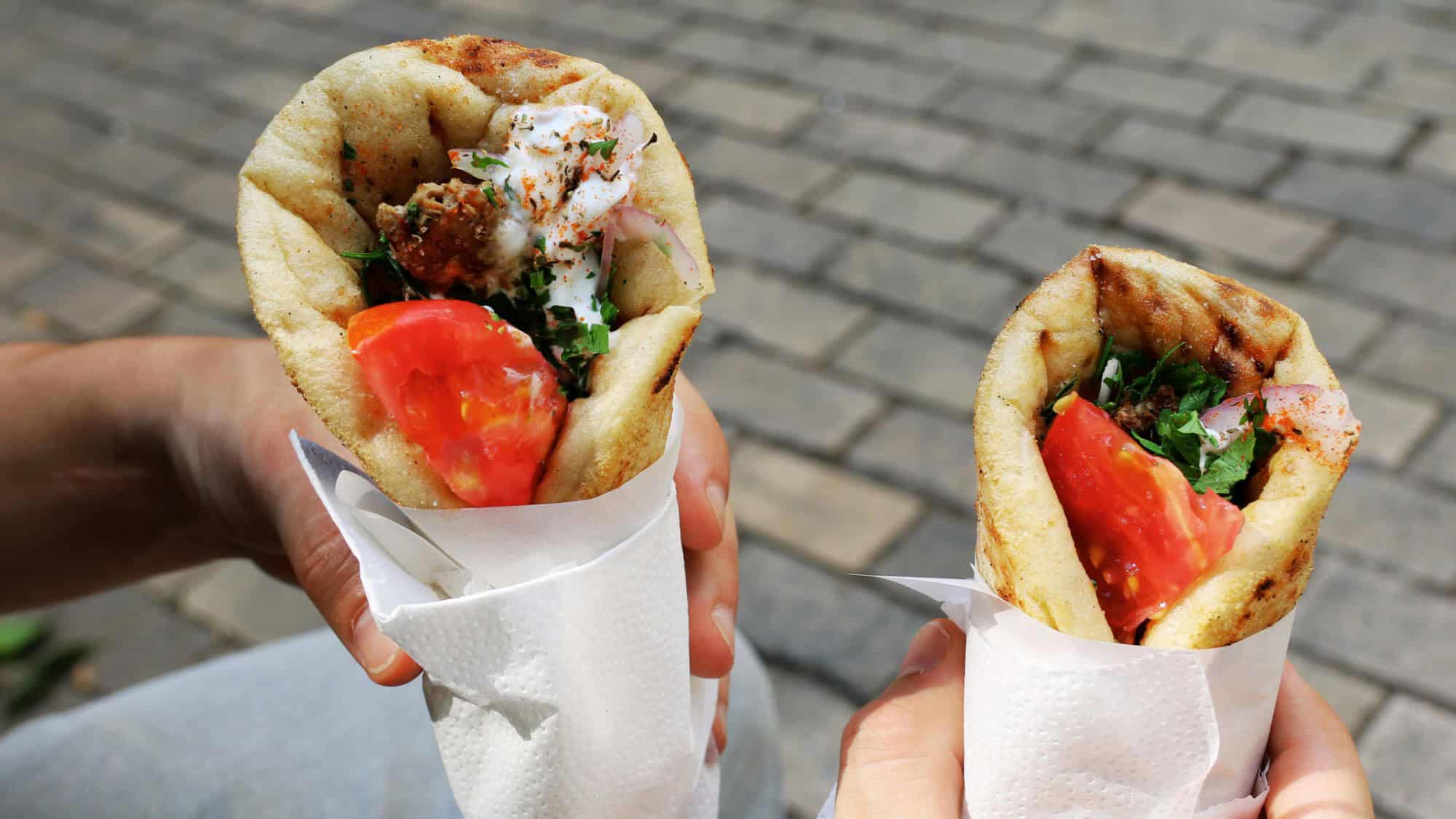 Two hands hold warm pita wraps stuffed with falafel, fresh vegetables, and creamy sauce.

