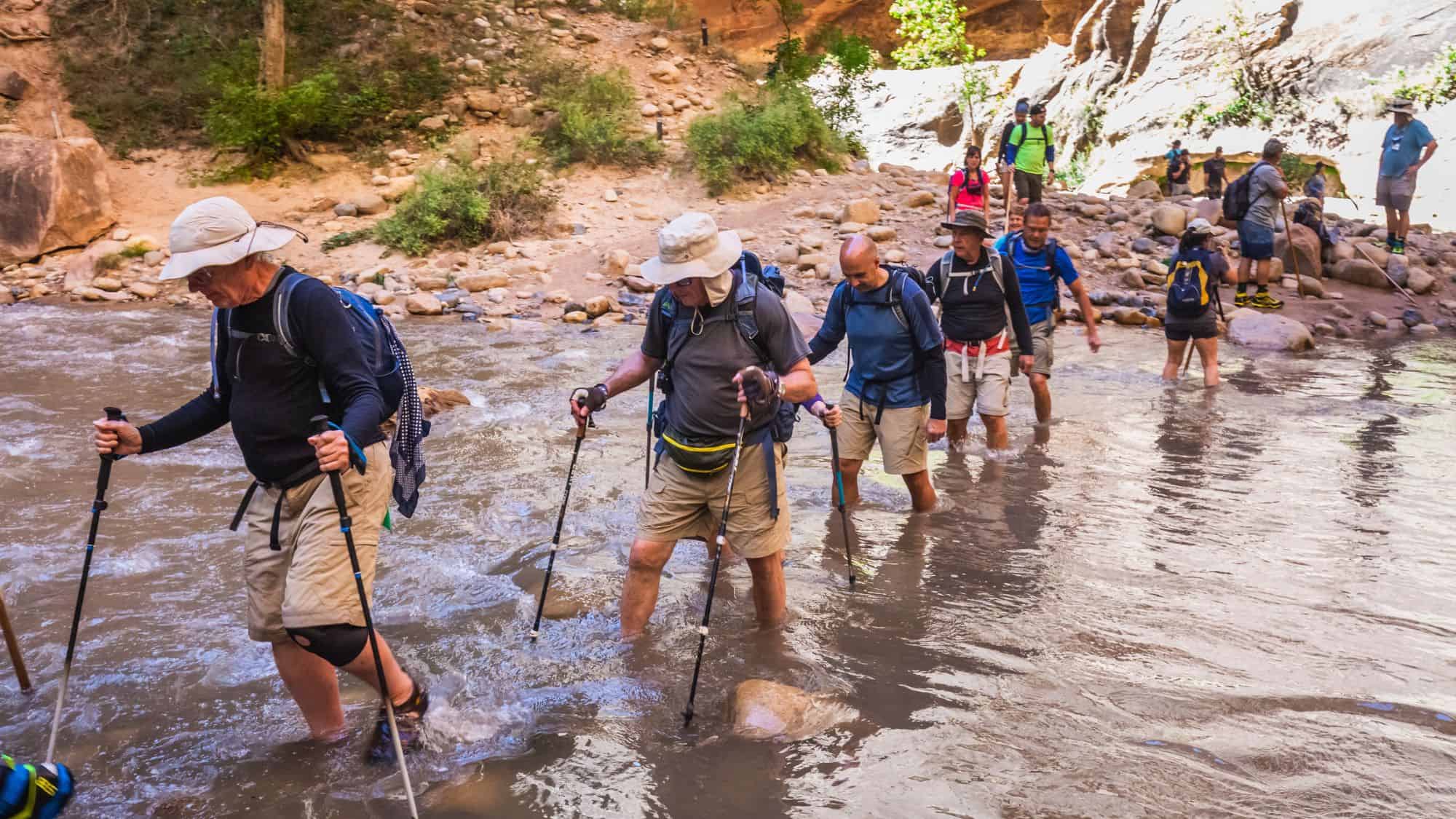 A group of hikers wade through a shallow rocky river in a canyon, using walking sticks for balance.
