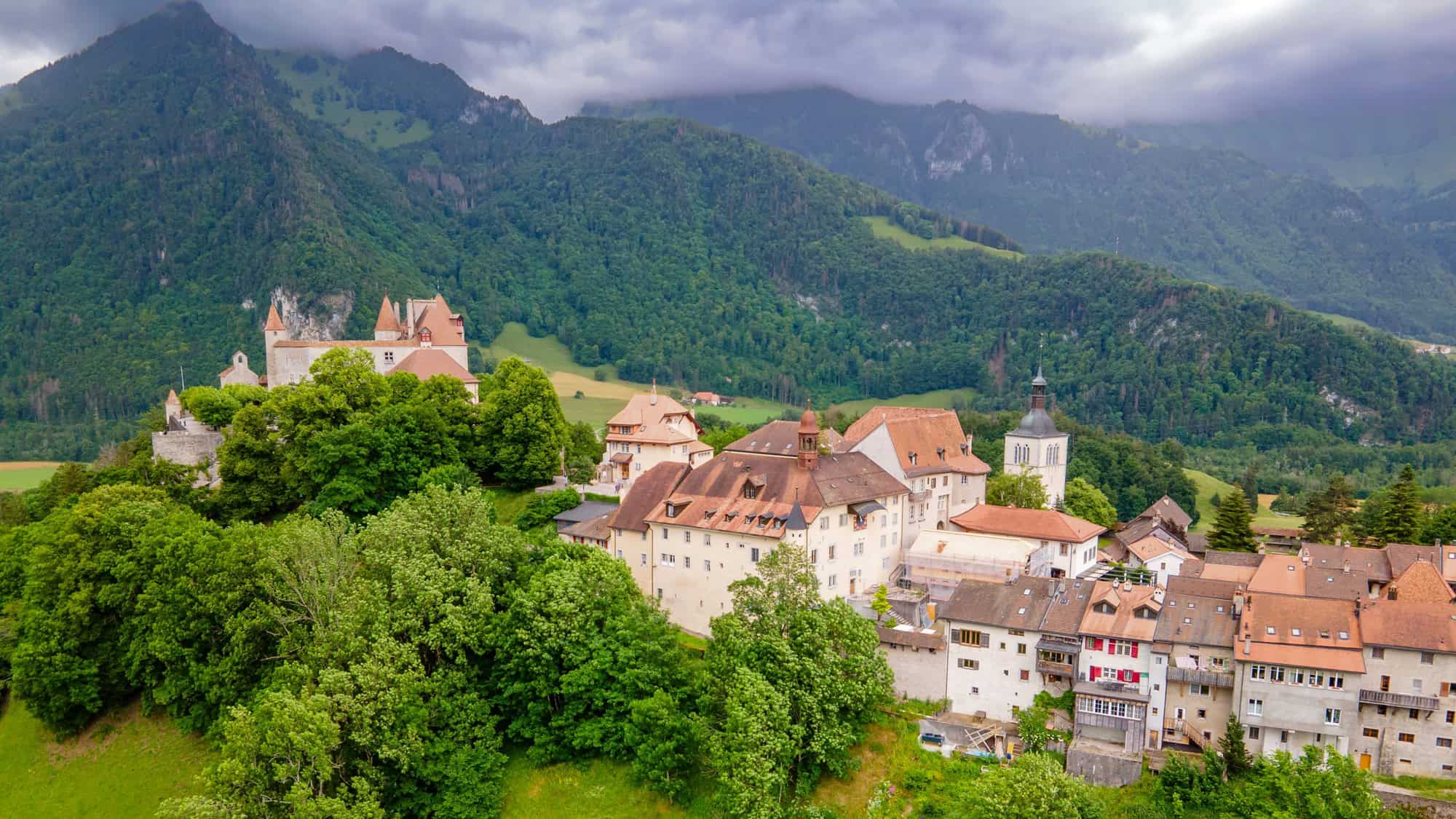A medieval hilltop village with terracotta rooftops and a castle is surrounded by dense green forests and mountains.