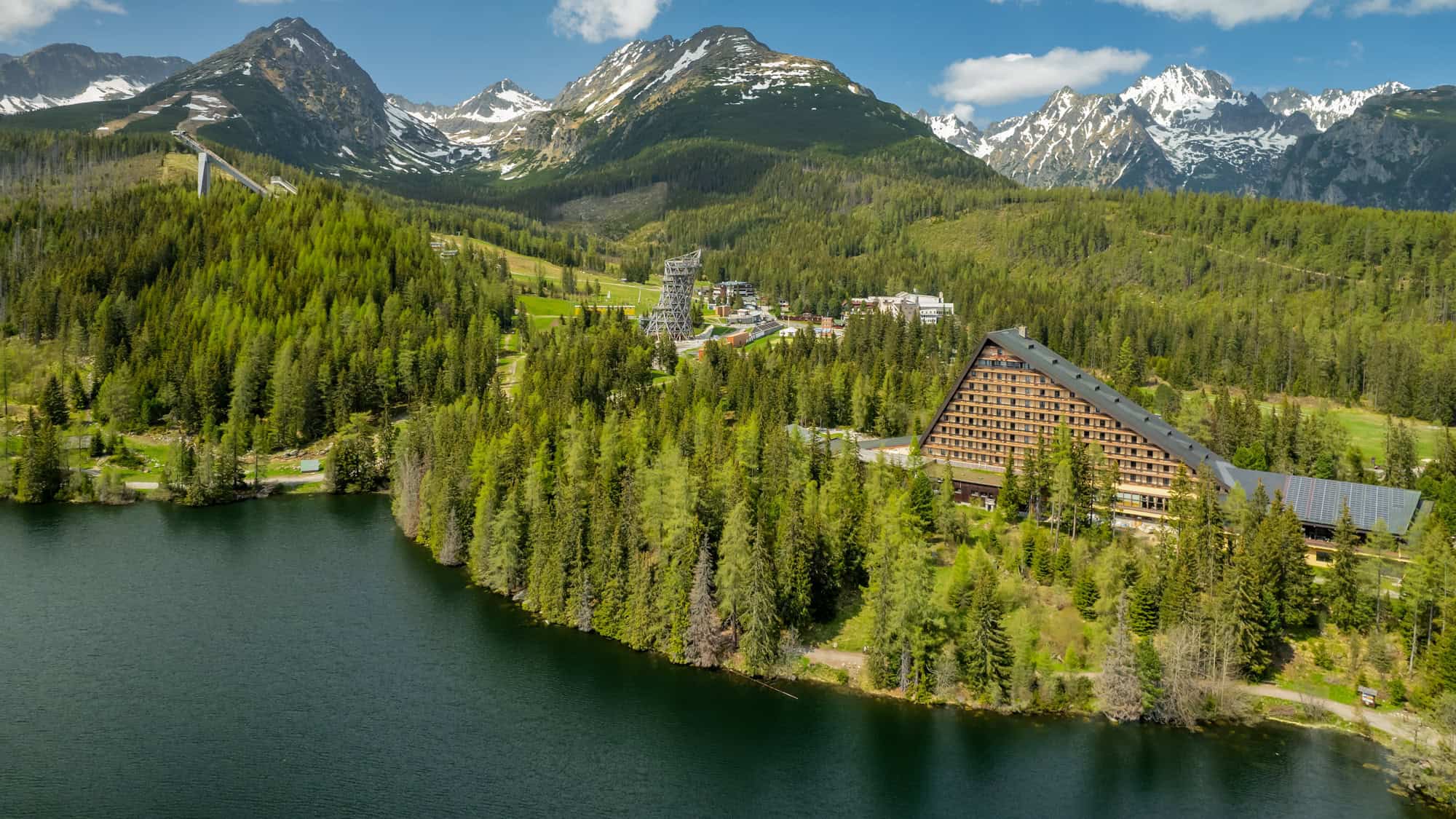 A large triangular resort building sits nestled among pine trees on the edge of a clear alpine lake, with snow-capped mountains towering in the background.
