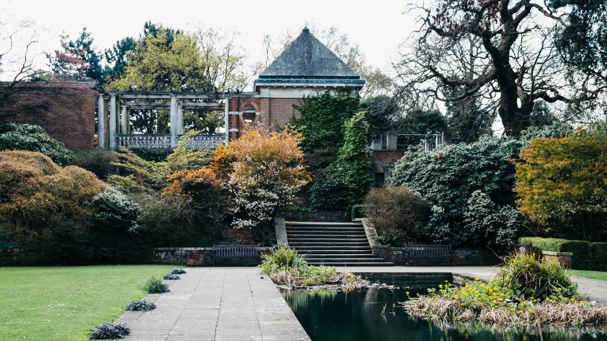 A landscaped garden with stone steps, lush foliage, and a reflecting pond leads up to a brick building draped in ivy.