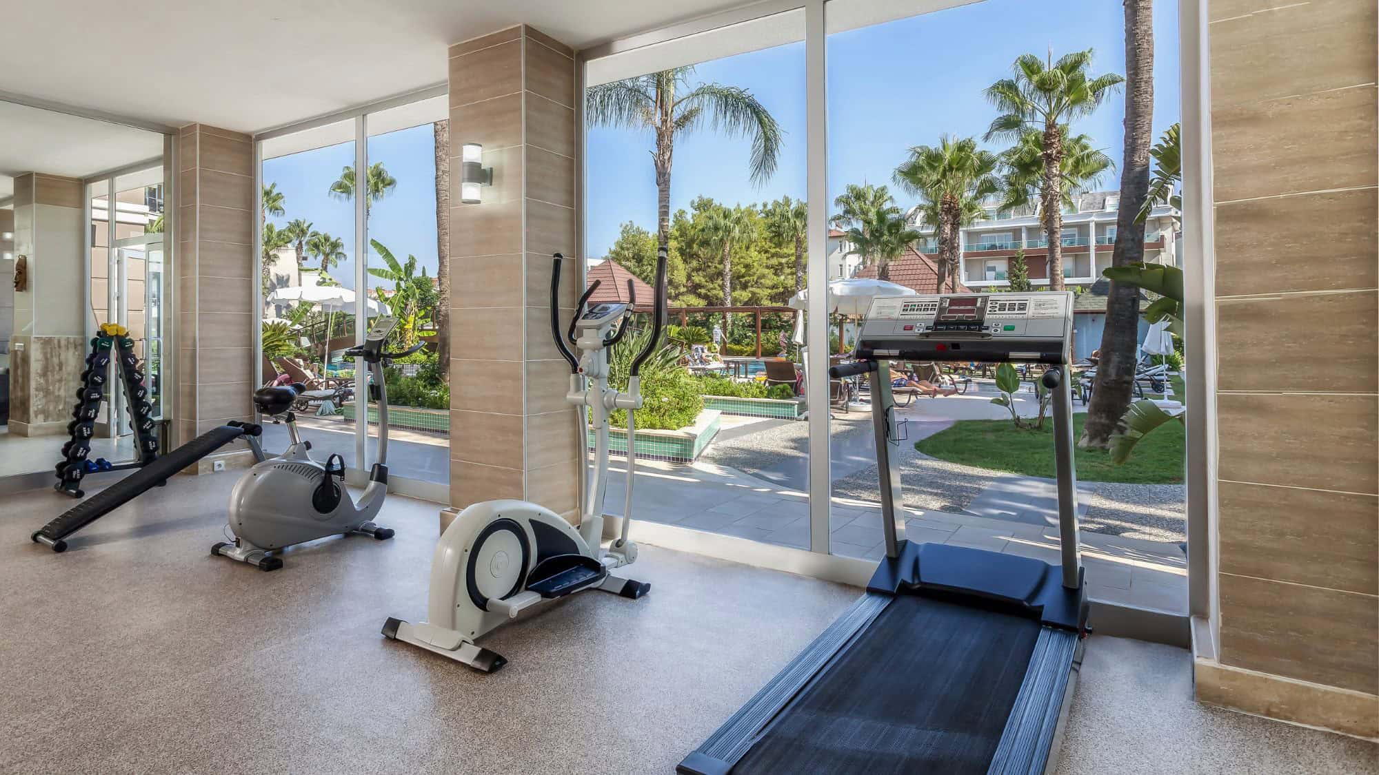 A bright, modern gym features cardio equipment and dumbbells beside floor-to-ceiling windows overlooking palm trees and a sunny resort courtyard.