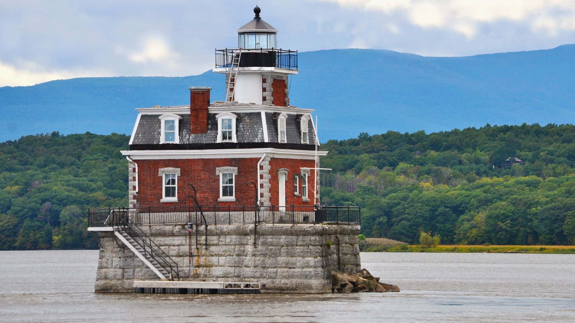 A red-brick lighthouse with a black lantern room stands on a stone base in the middle of the Hudson River, framed by forested hills and distant blue mountains.