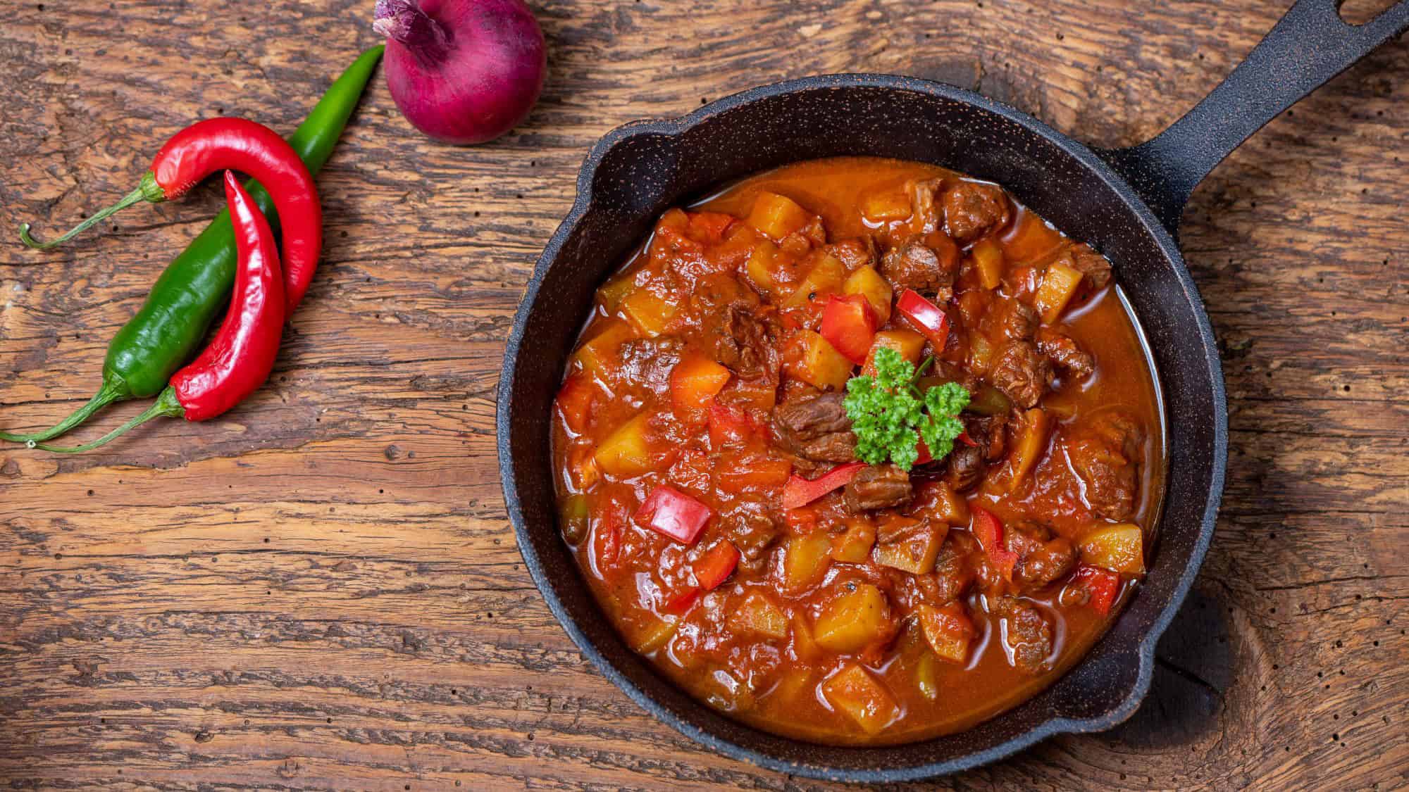 A rustic cast iron pan holds rich beef stew with chunks of potatoes, peppers, and tomatoes, garnished with parsley.