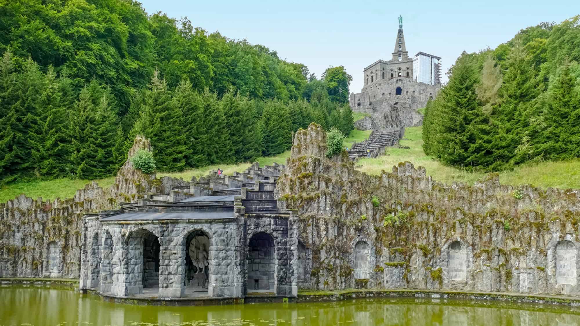 Stone arches and terraces lead up to the grand Hercules monument, set against a backdrop of dense forest.
