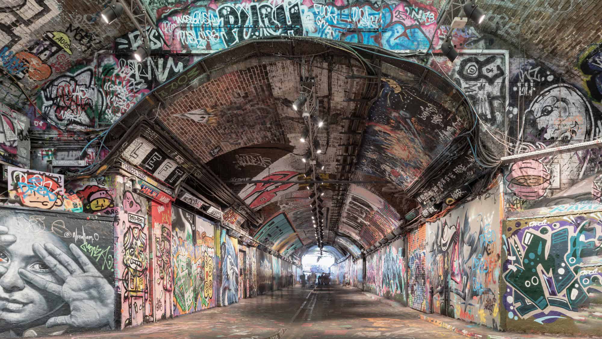 A brick arch tunnel is completely covered in colorful graffiti and street art with spotlights illuminating the walls and ceiling.