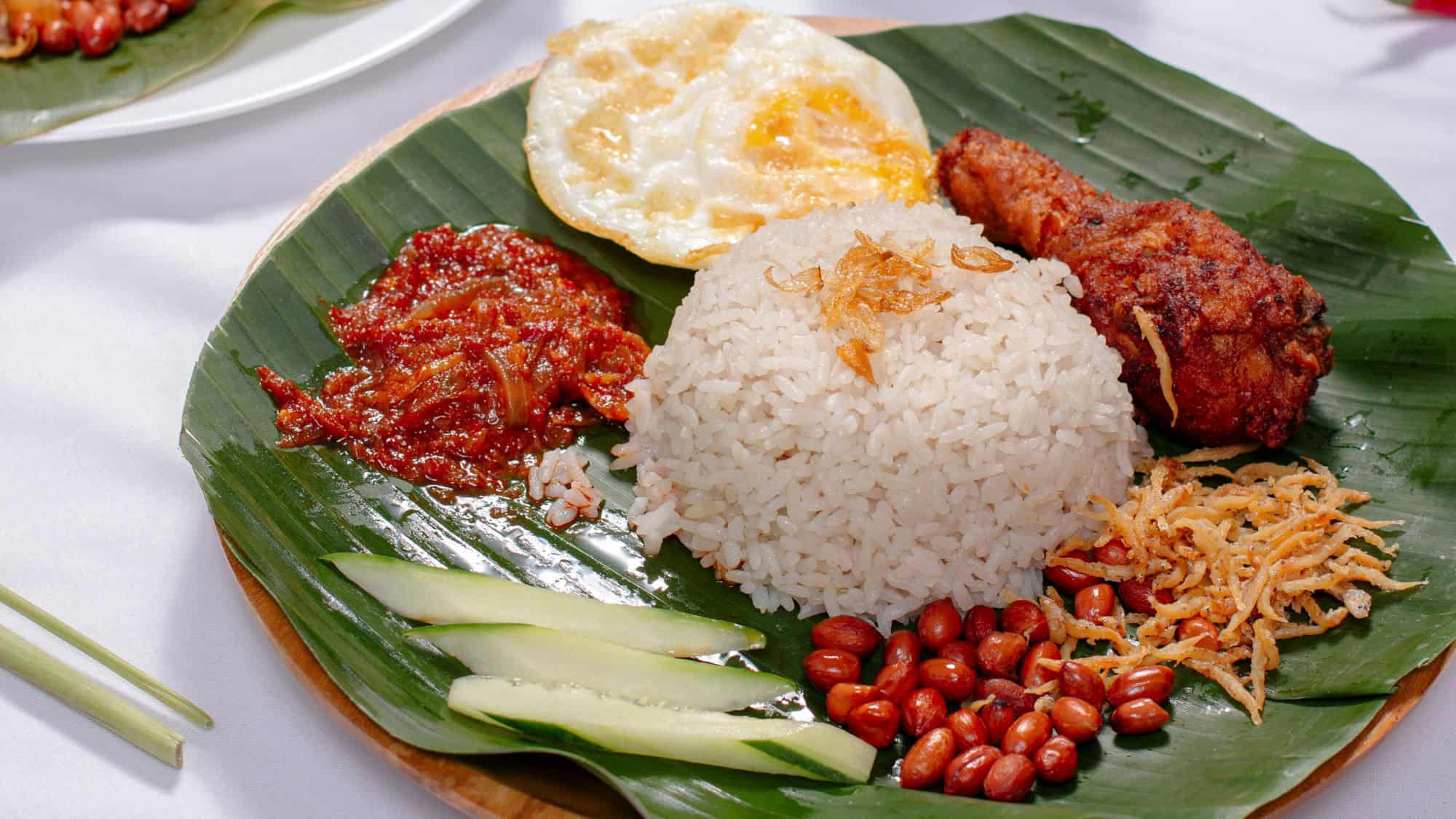 A banana leaf-lined plate holds coconut rice, fried chicken, sambal, cucumber slices, peanuts, and a fried egg.