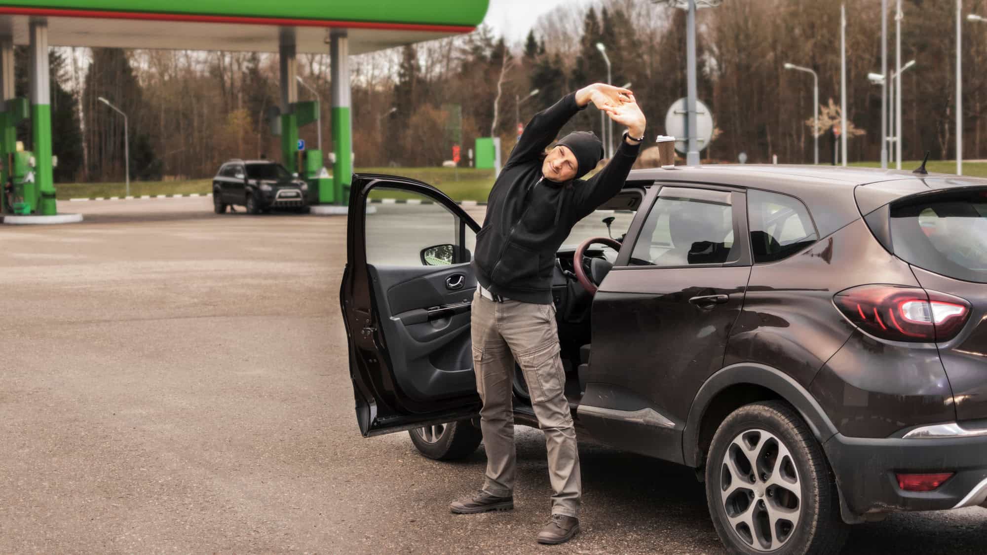 A man in casual outdoor clothing stretches beside his parked car at a gas station, with forested surroundings and fueling pumps in the background.