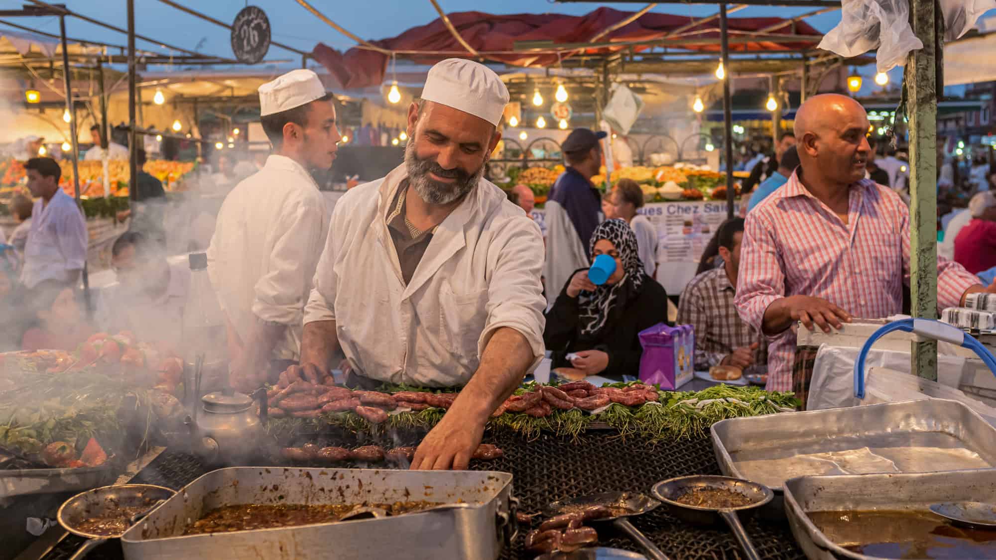A vendor in white attire grills skewers of meat at a bustling open-air night market.