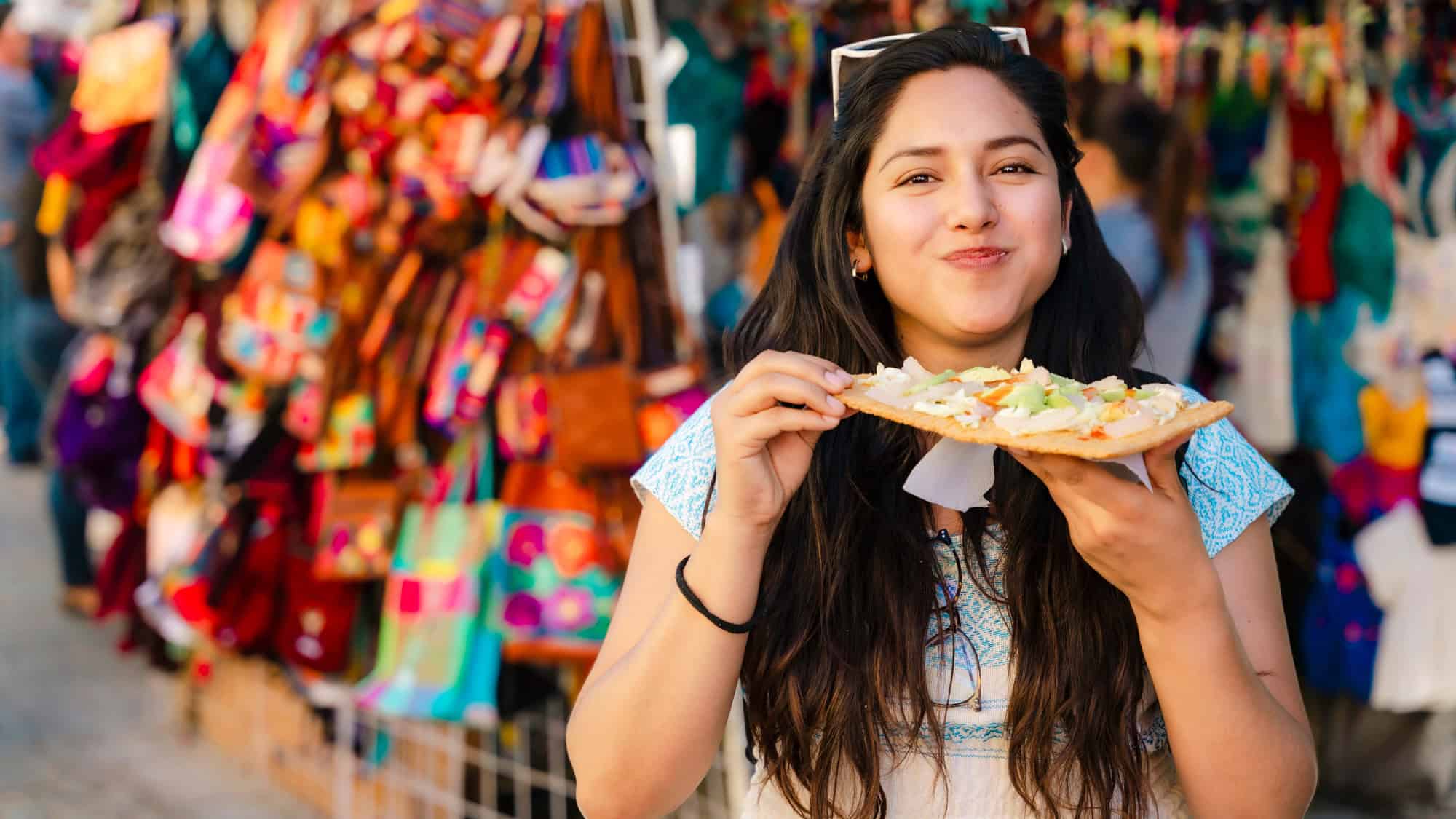 A smiling woman enjoys a crispy tostada topped with fresh vegetables at a colorful outdoor market.