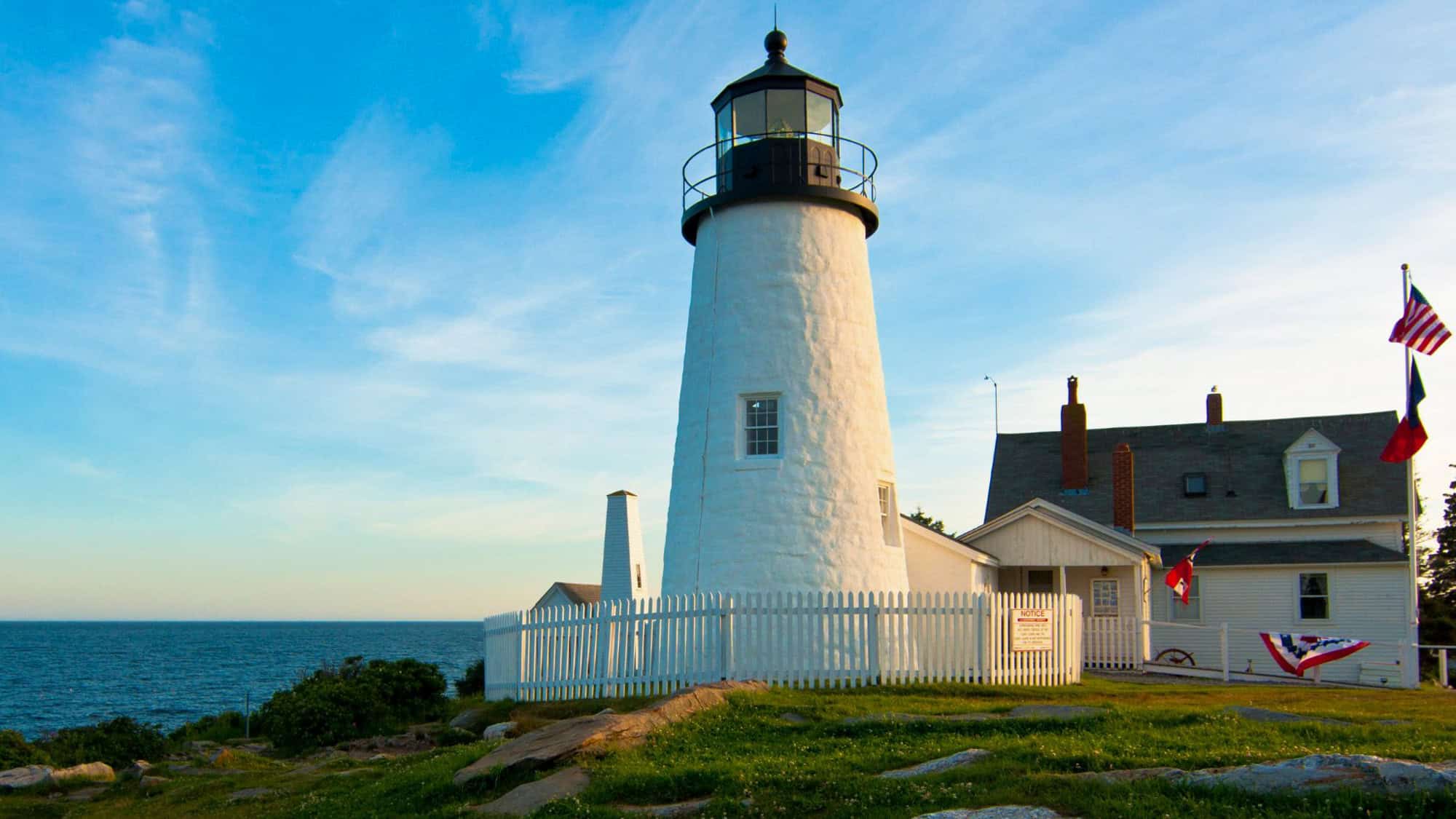 The iconic white tower of Pemaquid Point Lighthouse rises above a rocky coastline, bordered by a white picket fence.