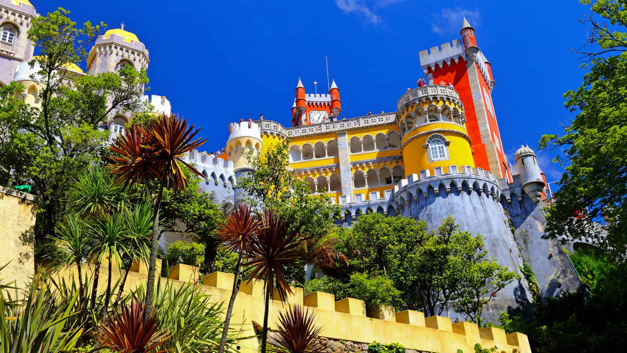 The vibrant red, yellow, and gray towers of Pena Palace rise above lush gardens under a bright blue sky in Sintra.