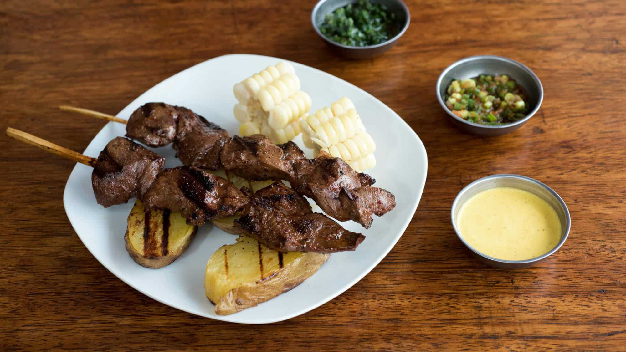 Two skewers of grilled beef heart rest on roasted potato slices and corn, served with small bowls of sauces and salsa.