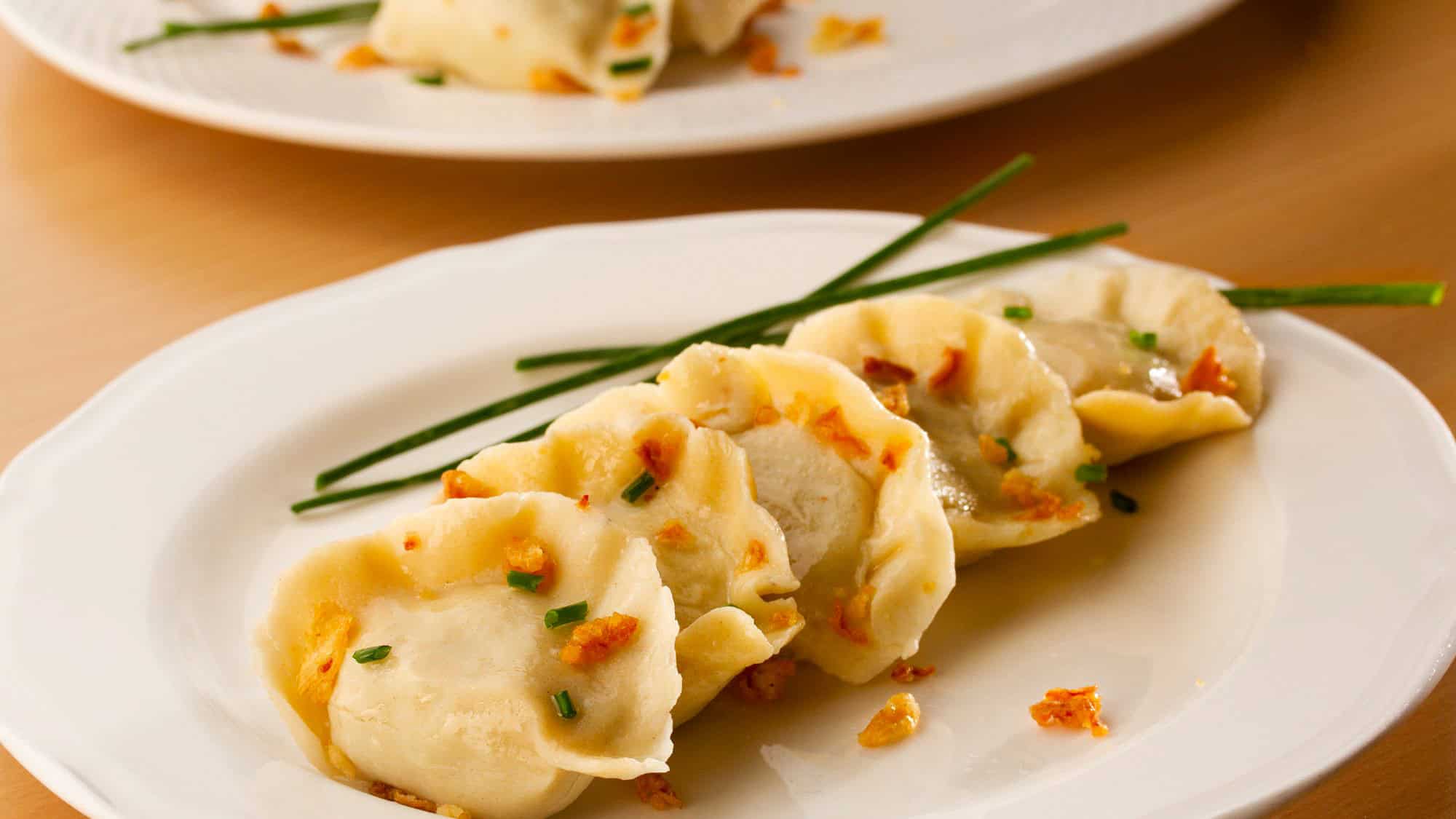 A row of golden-topped dumplings garnished with chopped chives and crispy bits sits on a white plate.