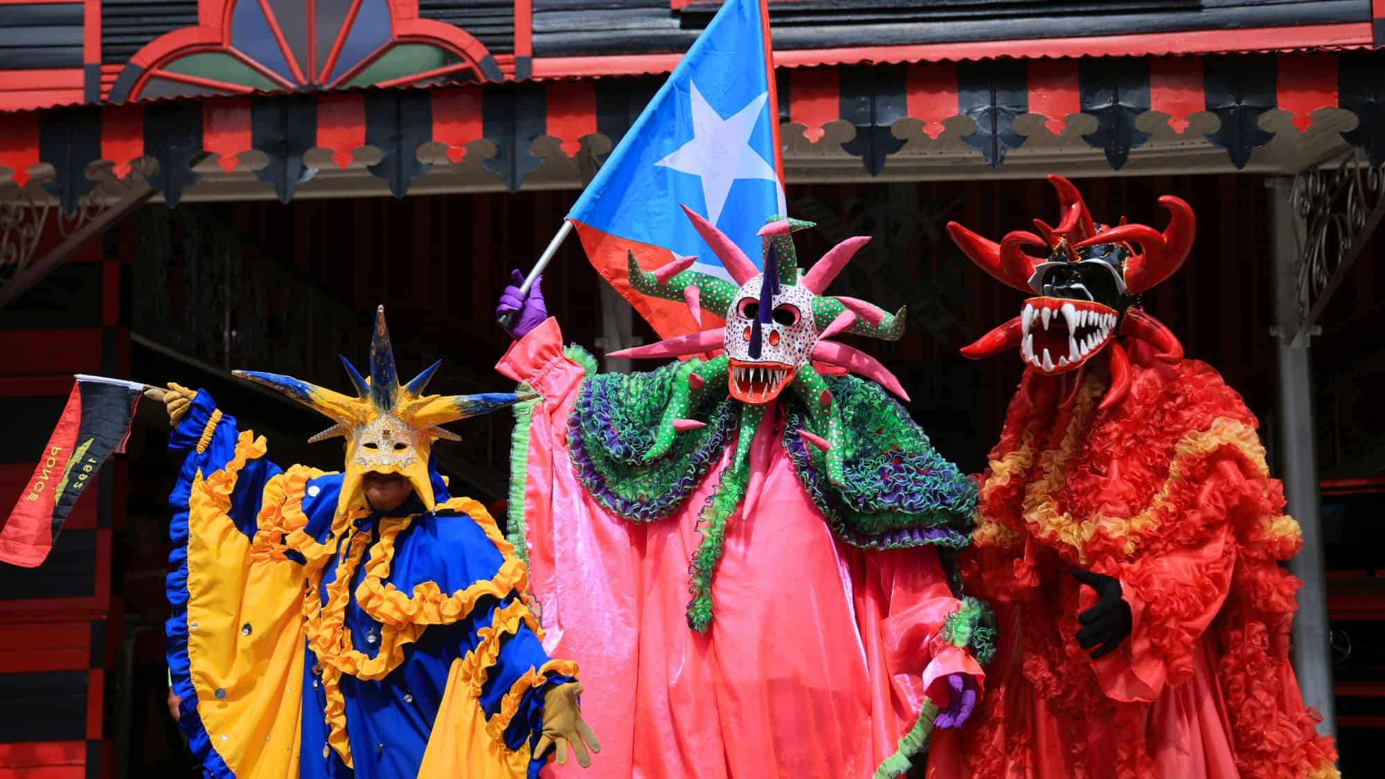 Three people in colorful, elaborate costumes and horned masks celebrate with a Puerto Rican flag during a festival.