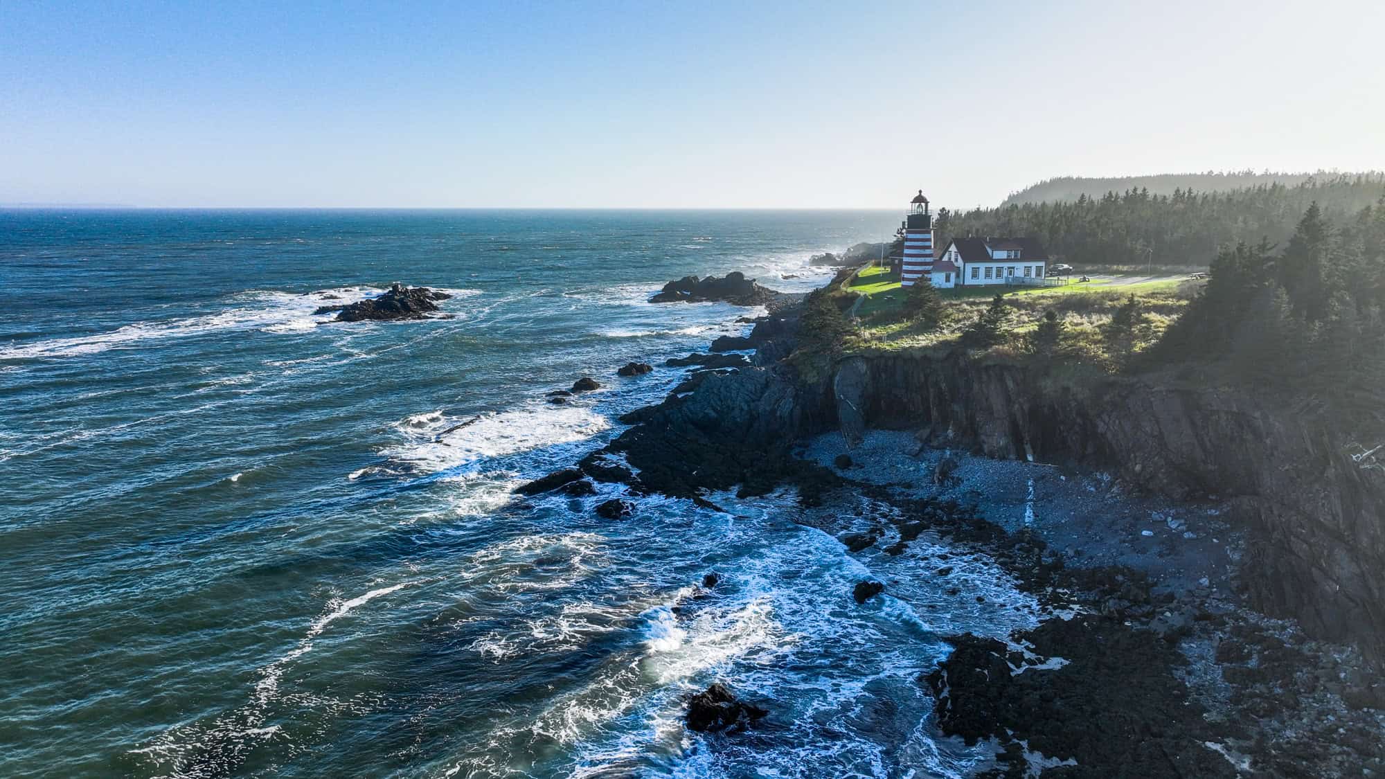 An aerial view of Maine’s red-and-white striped West Quoddy Head Lighthouse perched on a rocky cliff above crashing waves.