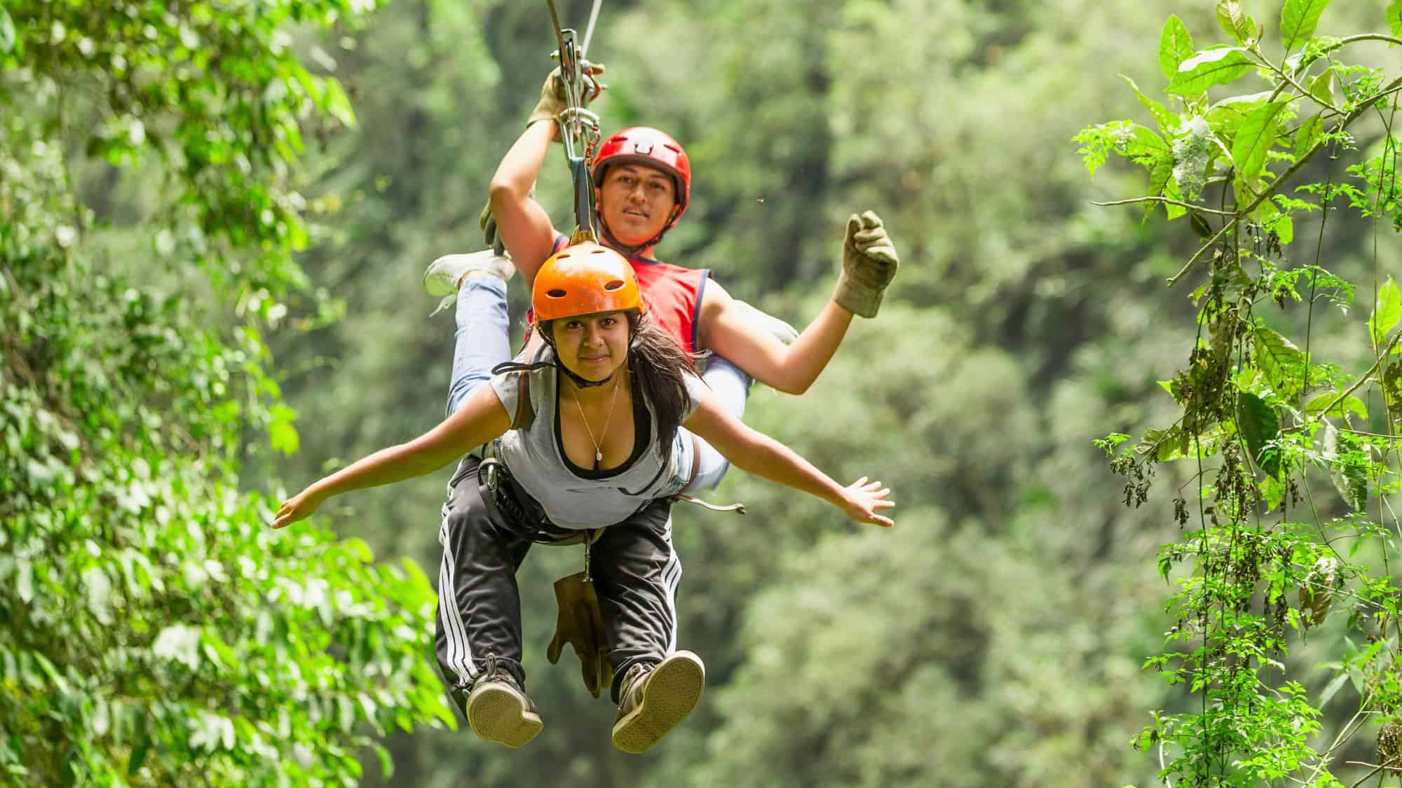 Two people wearing helmets glide together on a zipline through a lush tropical forest.