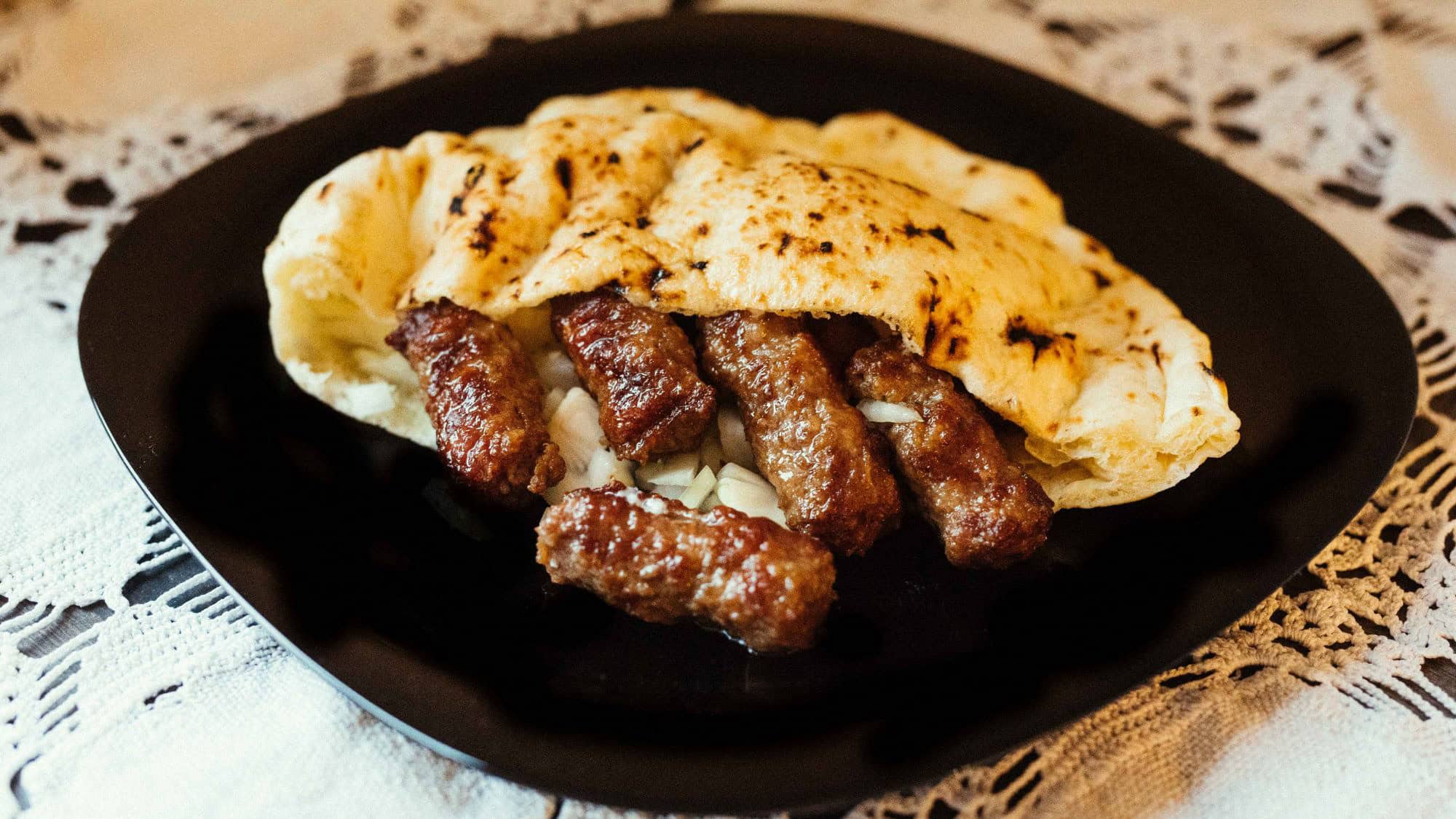Grilled Balkan sausages served inside a fluffy flatbread with chopped onions on a black plate.