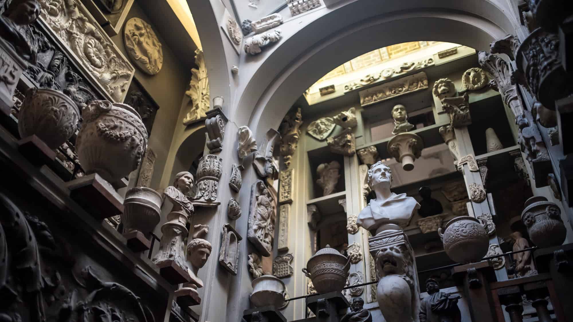 A high-ceilinged museum gallery is filled with plaster casts of classical busts, urns, and architectural fragments.