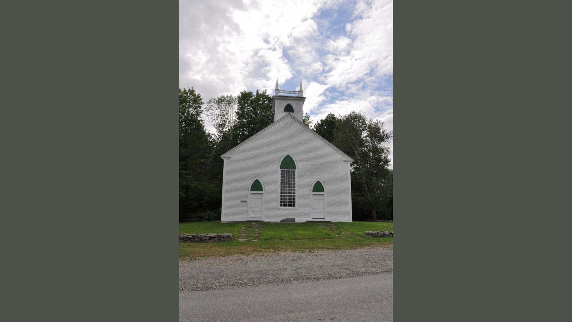 A simple white wooden church with green-trimmed windows and twin spires stands in front of a backdrop of trees under a partly cloudy sky.