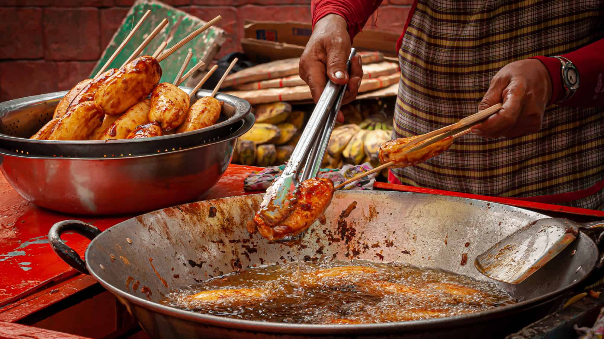 A vendor uses tongs to lift caramelized skewered bananas from a bubbling wok of hot oil, with a bowl of freshly fried bananas beside them.