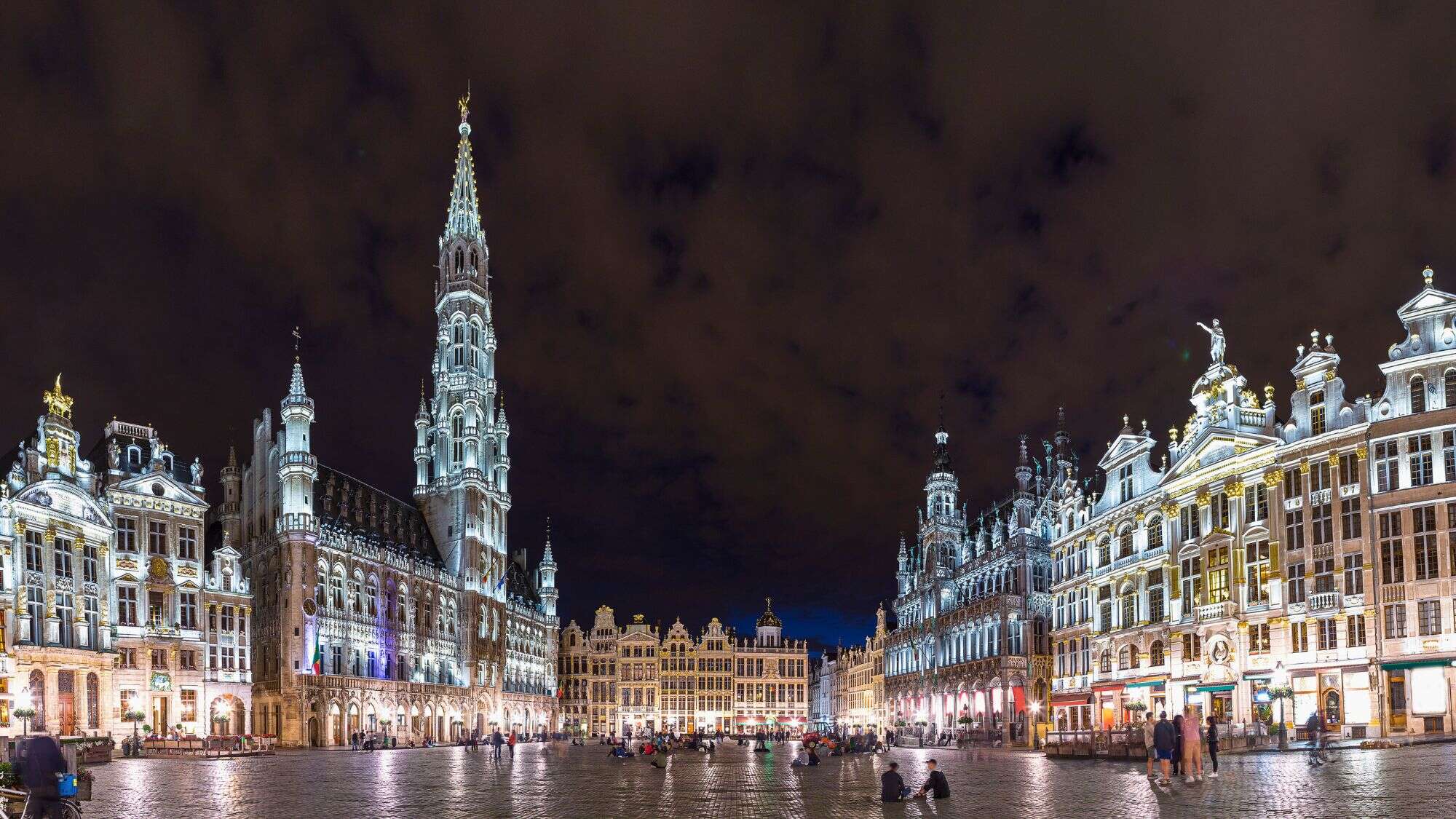 Brussels' Grand Place glows under dramatic night lighting, with ornate guildhalls and the towering Town Hall illuminated against a dark, clouded sky.
