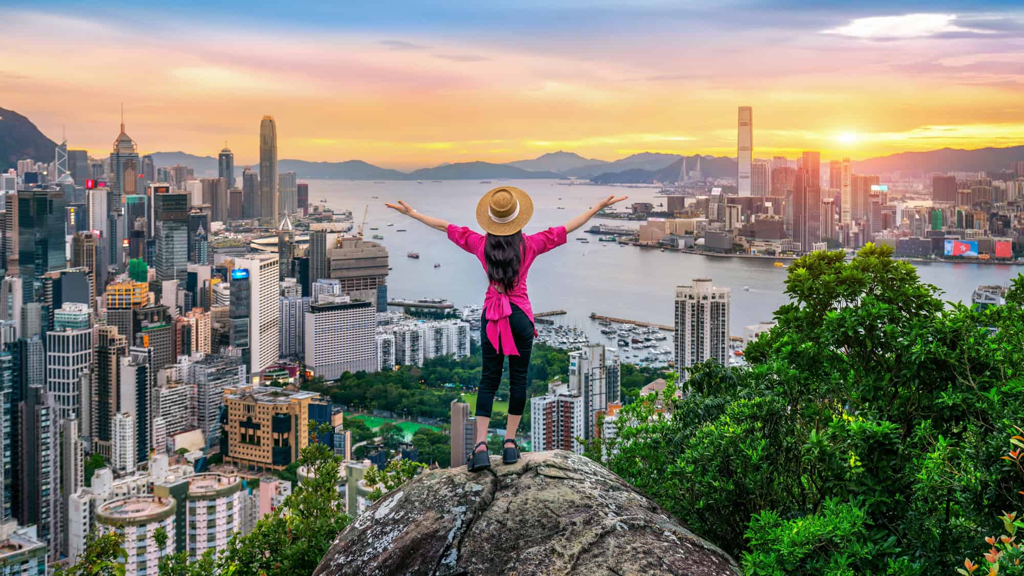 A woman in a straw hat and bright pink shirt stands on a rock with her arms raised, overlooking a sprawling city skyline at sunset from a lush hilltop.