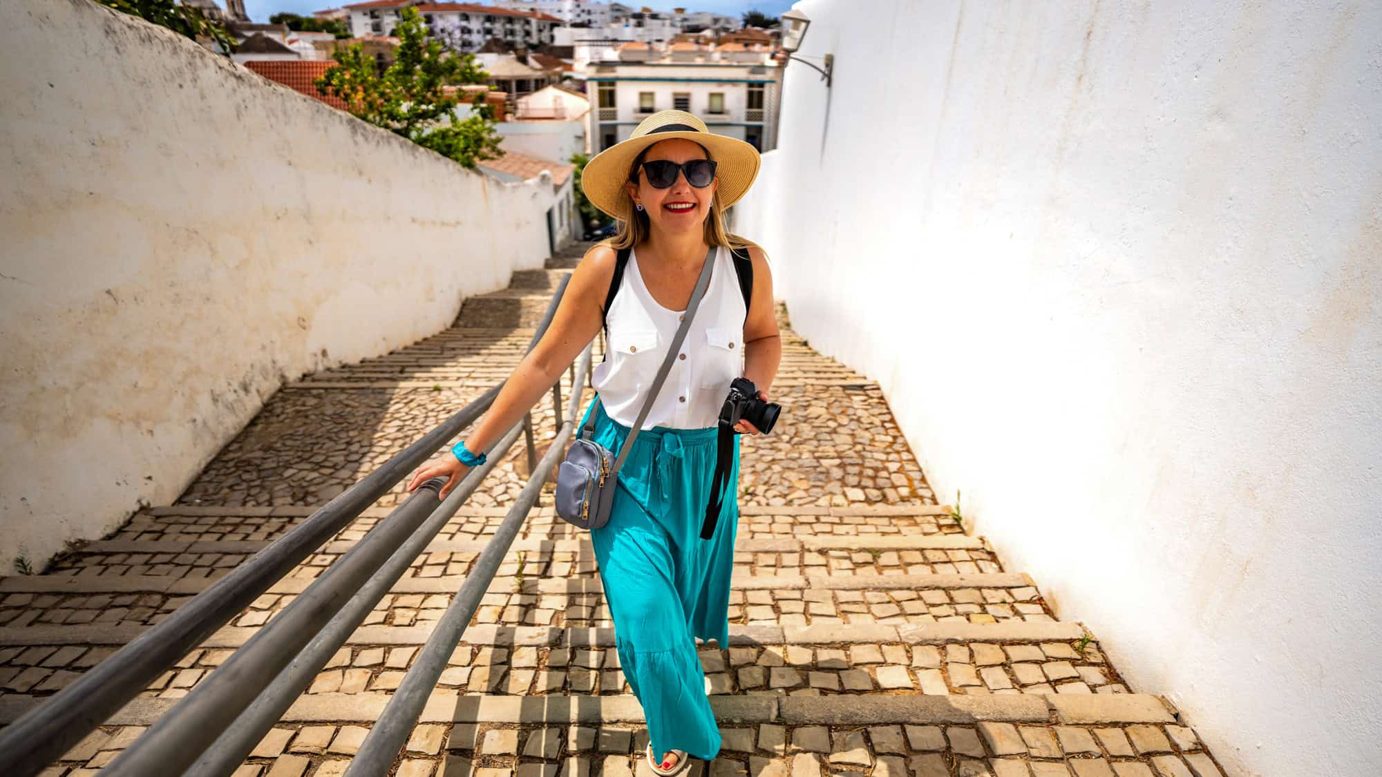 A smiling woman in a sunhat and turquoise skirt walks up a narrow cobblestone staircase between whitewashed walls, holding a camera and enjoying a sunny day.
