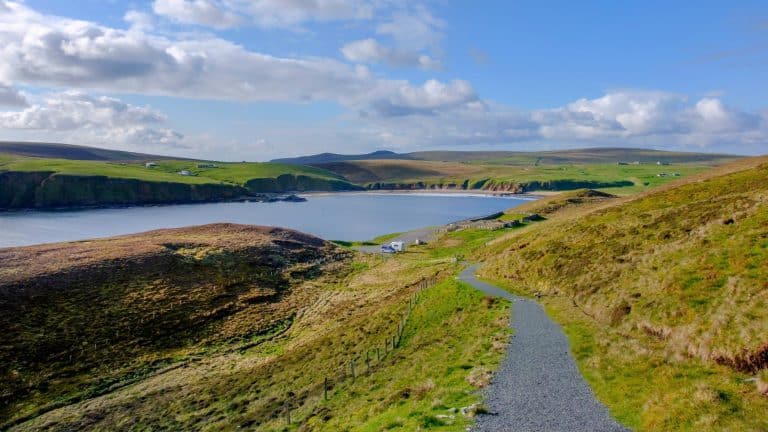 A winding path leads down a grassy hillside to a quiet bay, with gentle cliffs and scattered rural buildings under a partly cloudy blue sky in the Shetland Islands.