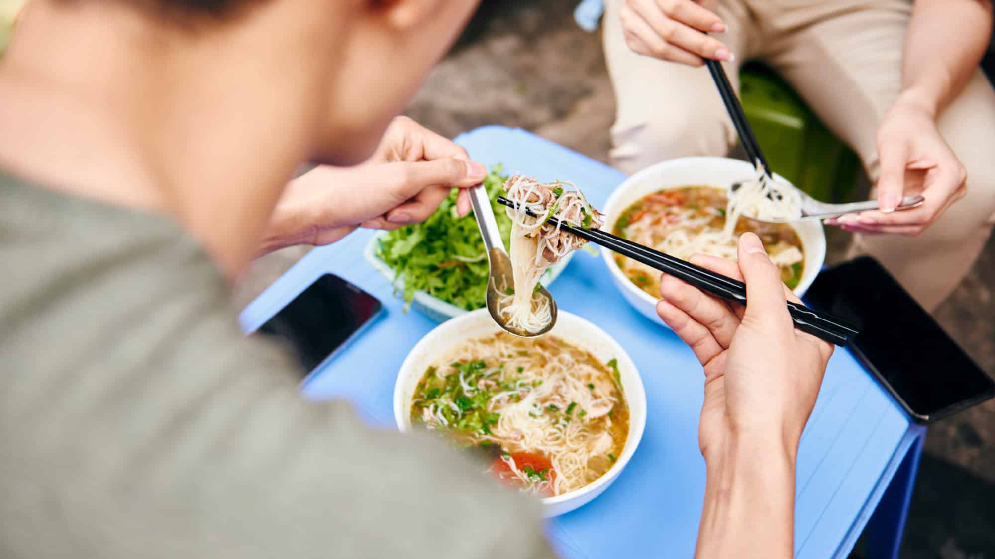 Two people use chopsticks and spoons to share steaming bowls of noodle soup at an outdoor table.